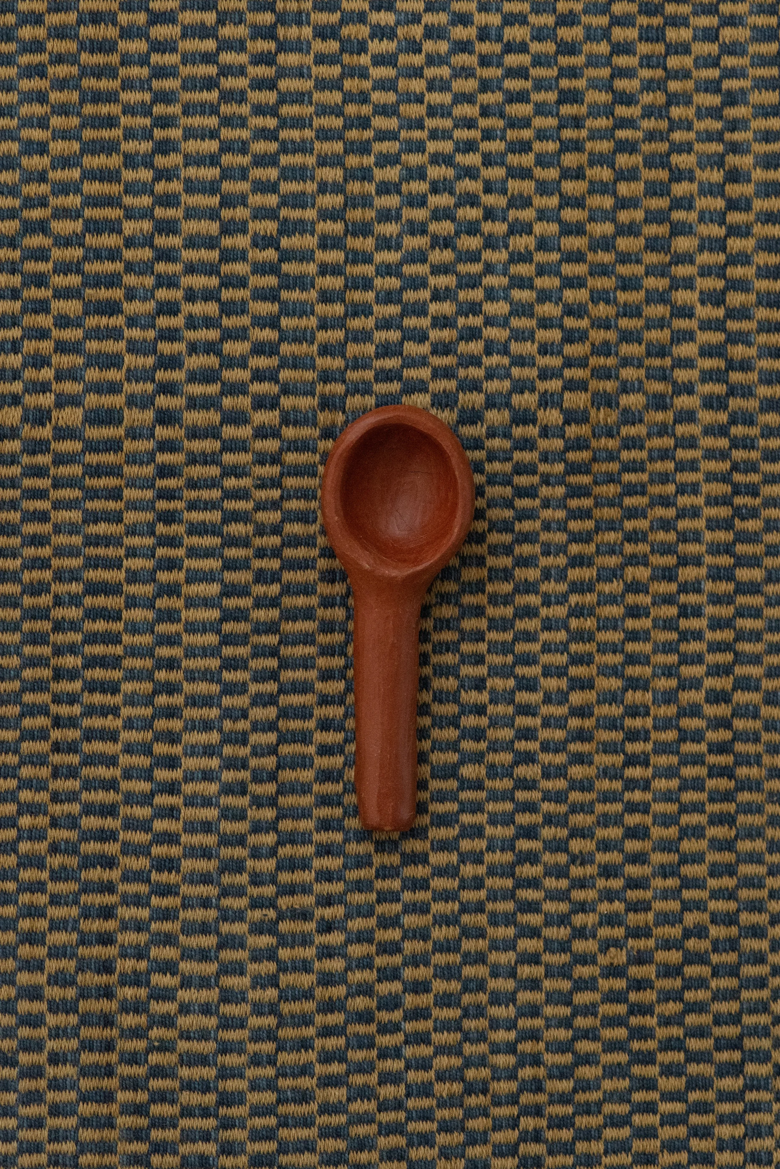 Top view of a Barro Rojo handmade red clay spoon from Oaxaca, placed on a woven textile background.