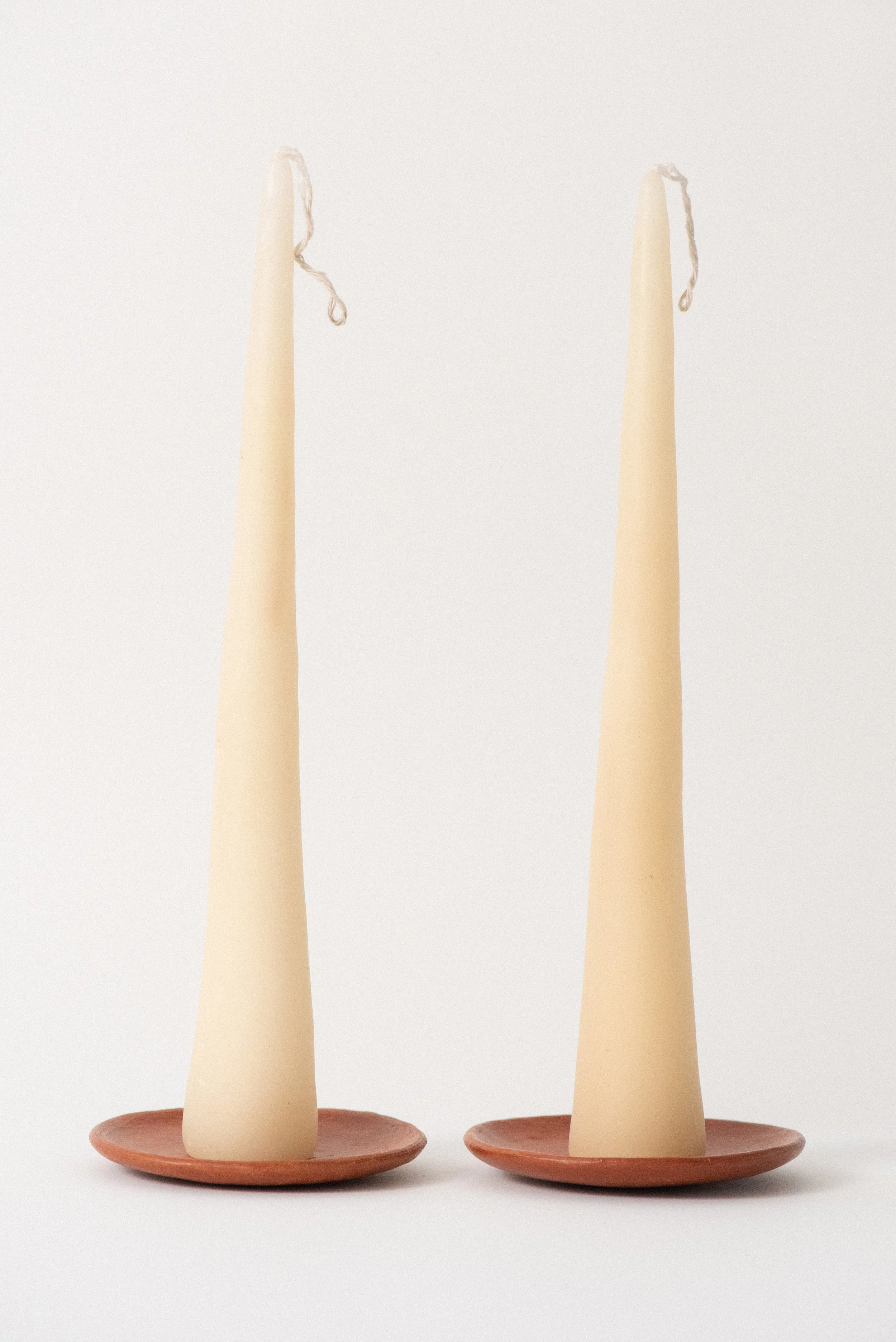 A pair of handmade beeswax taper candles from Oaxaca placed on barro rojo red clay plates against a white background.
