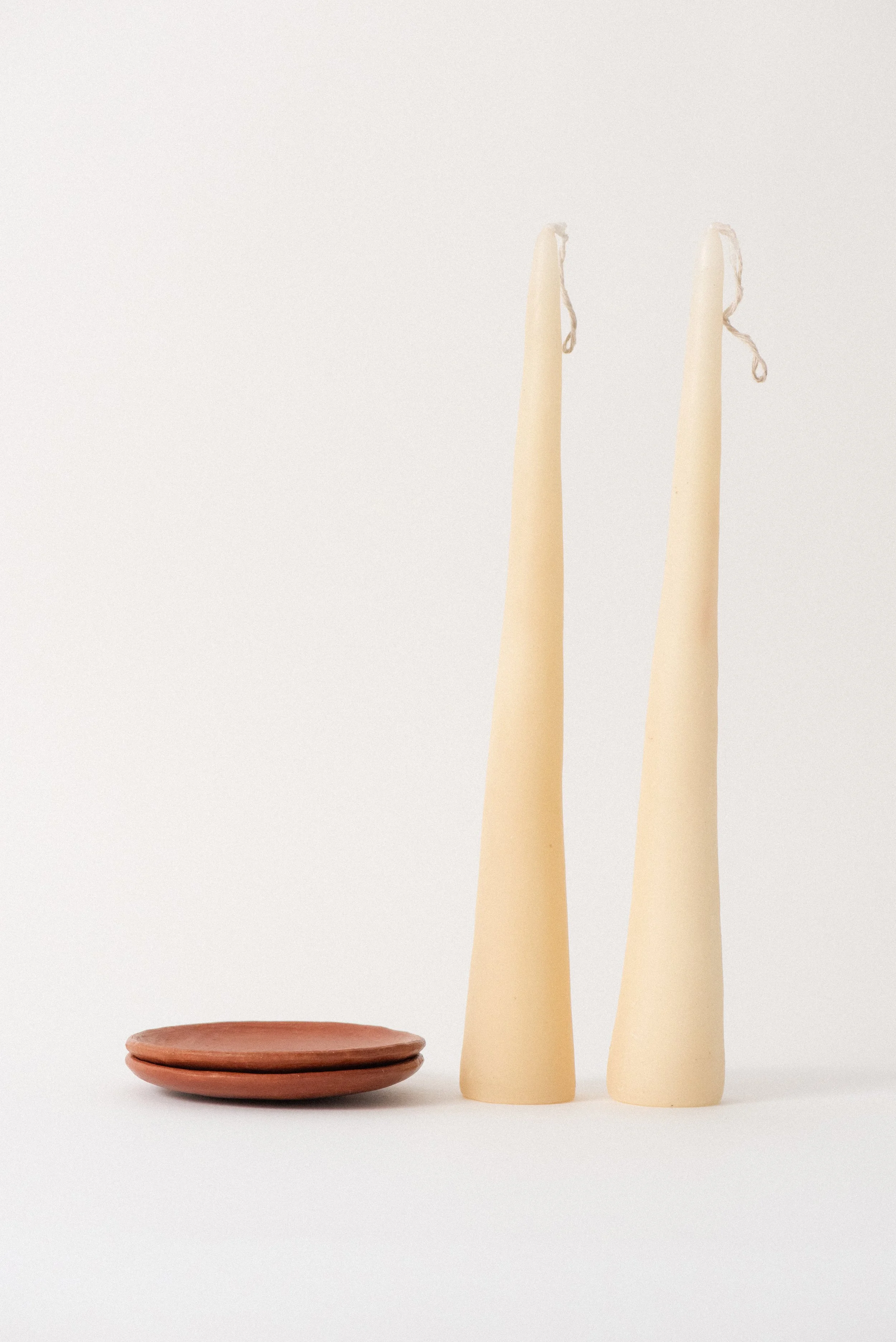 Beeswax taper candles from Oaxaca resting on handmade barro rojo red clay plates against a woven mat backdrop.