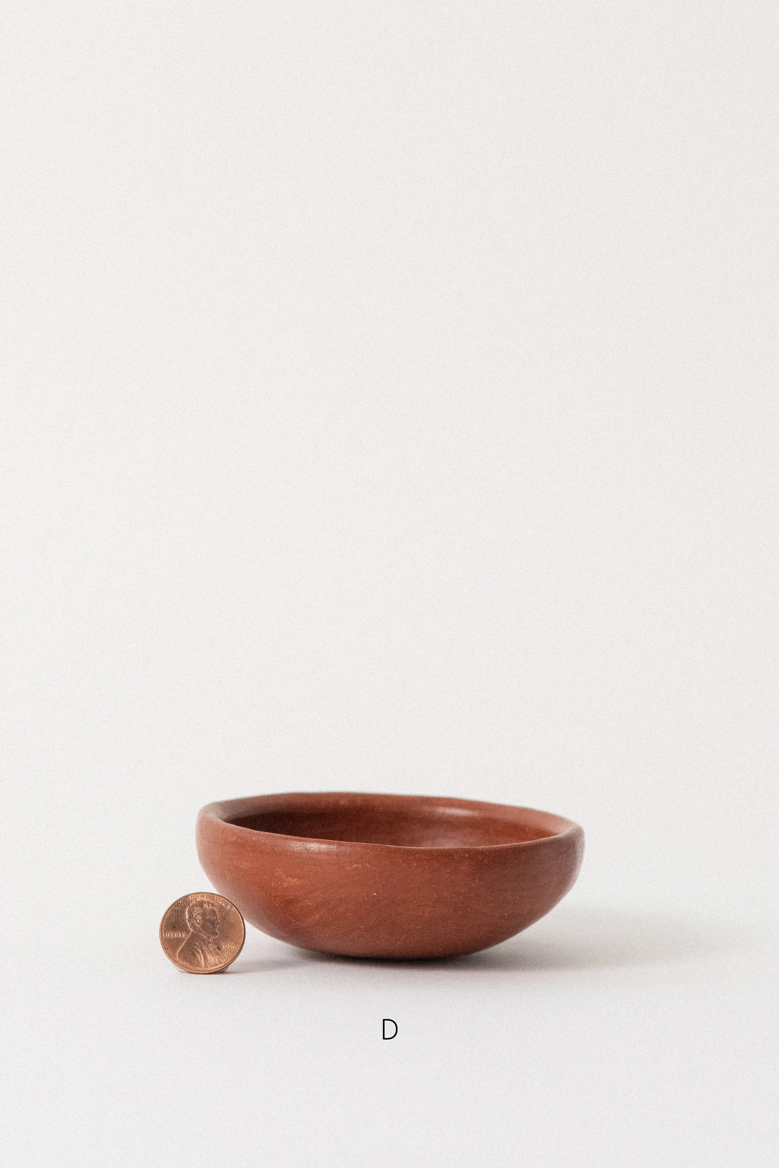 Single handmade barro rojo red clay bowl labeled D, crafted in Oaxaca, Mexico, shown with a penny for scale against a white background — rustic and versatile kitchenware.