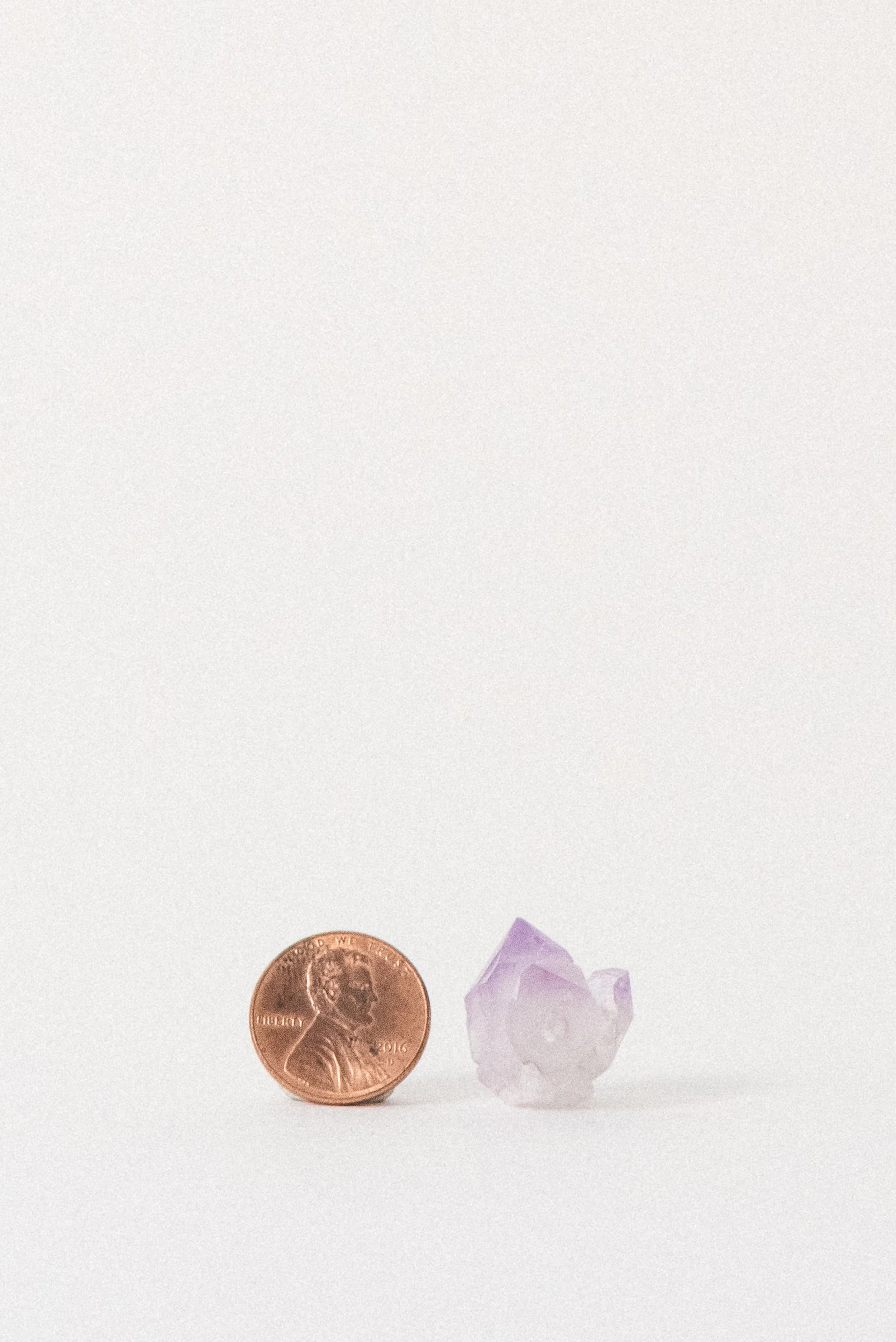 Small natural amethyst crystal from Jackson’s Crossroads, Georgia shown beside a U.S. penny for size reference; pale-to-violet point on white background