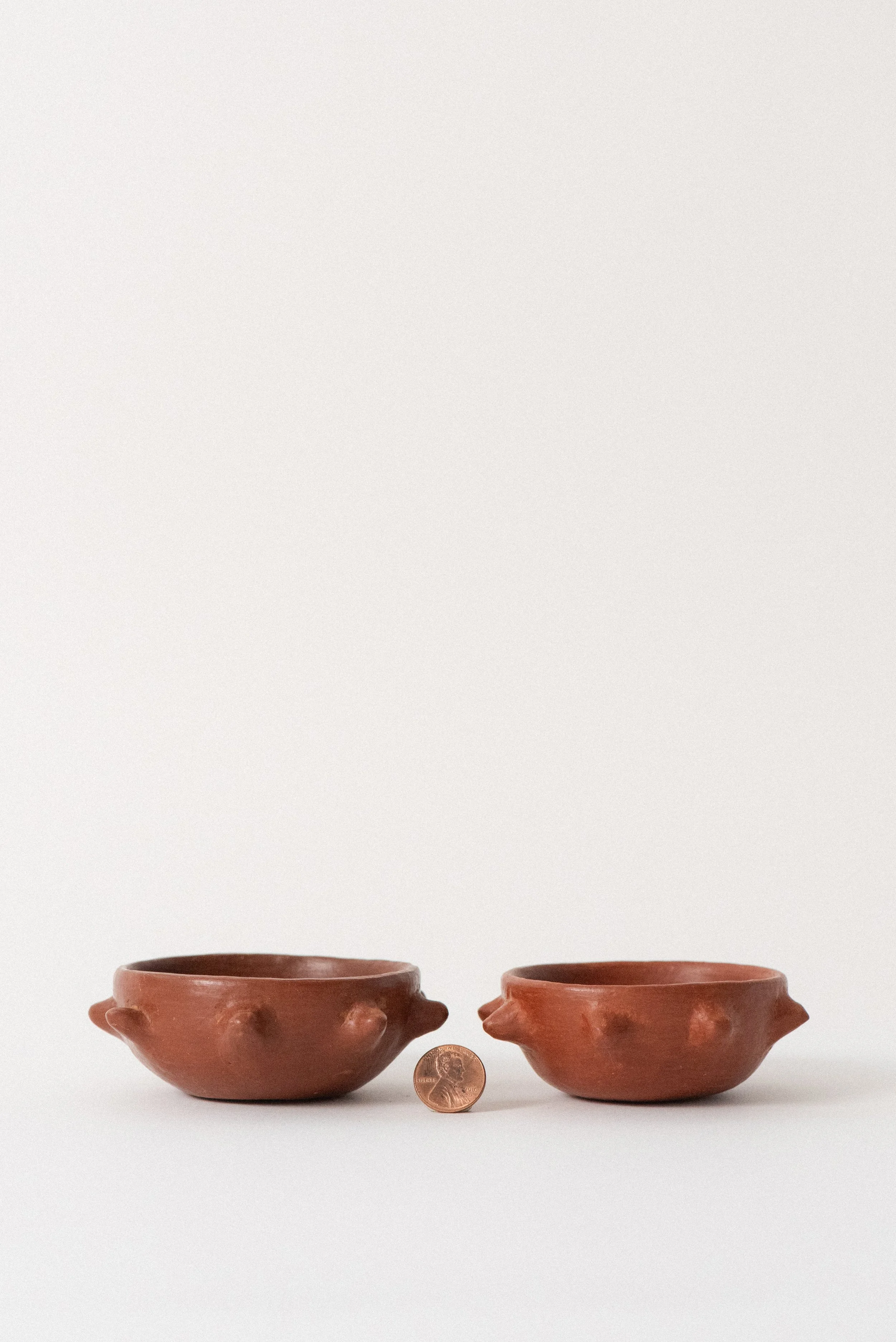 Two small red clay bowls shown with a penny for scale, highlighting the handmade size and organic shape of Zapotec pottery from Oaxaca.