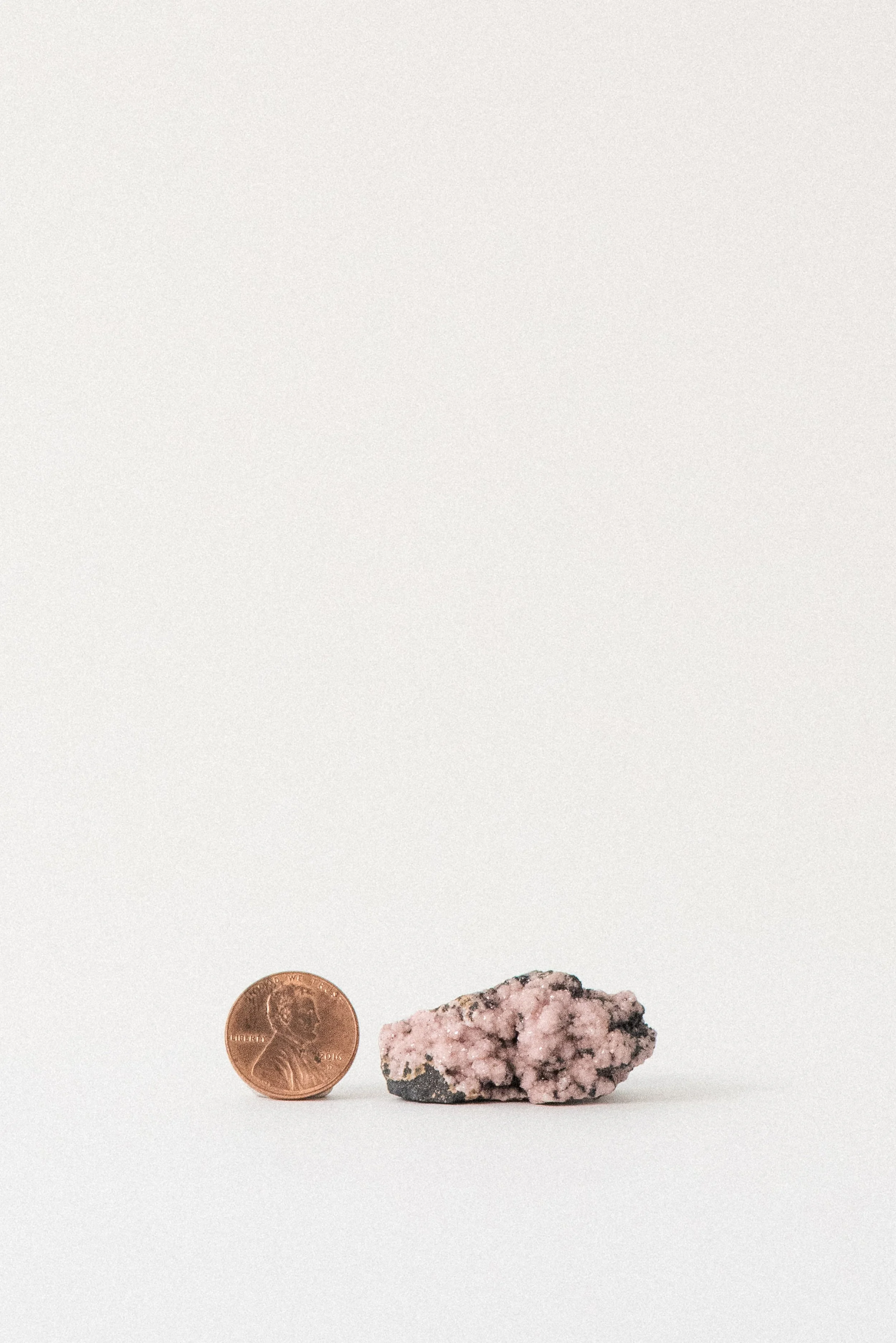 Pink rhodochrosite crystal from N’Chwaning Mine, South Africa, shown beside a U.S. penny for scale on a white background.
