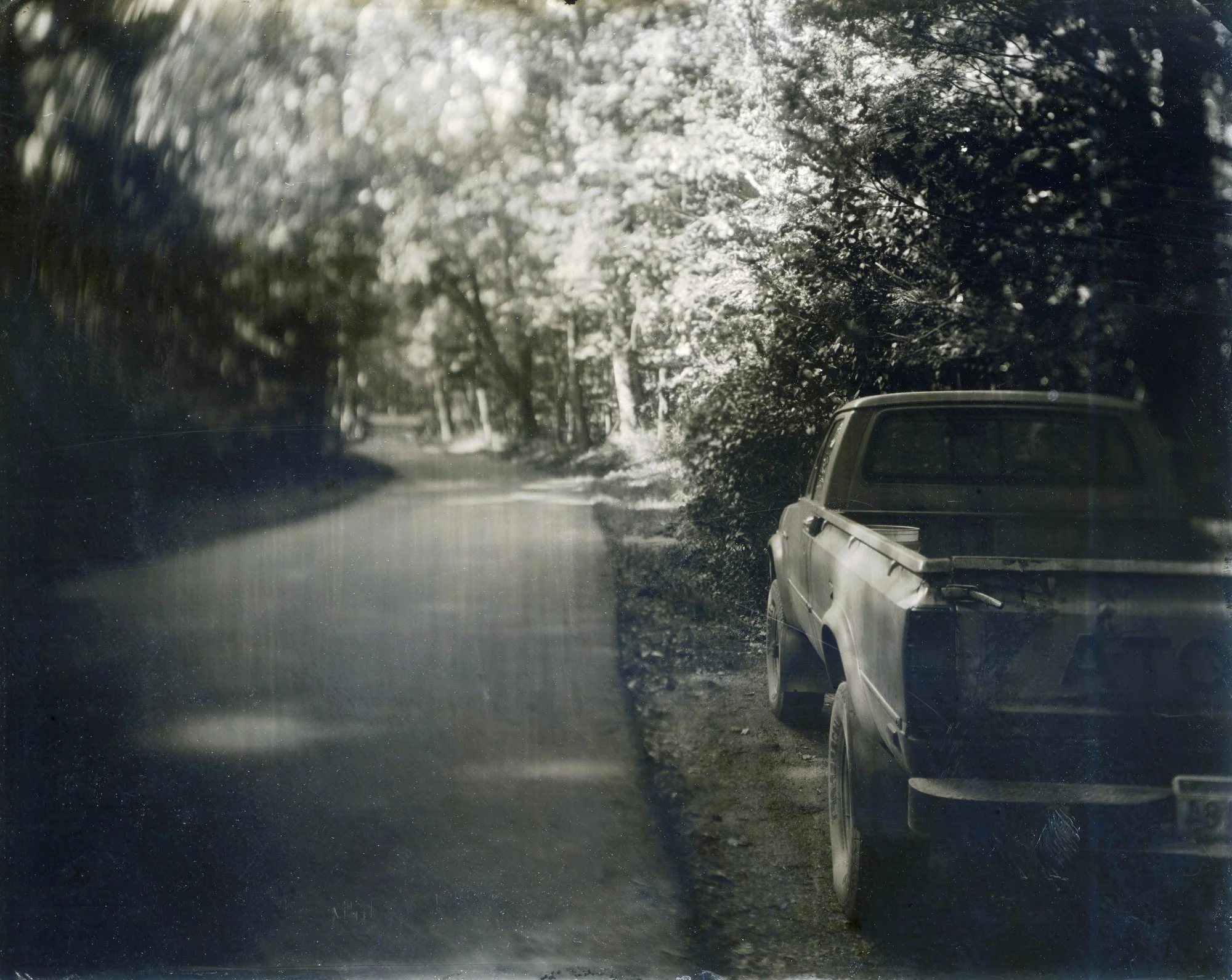 Tin type lanscape photo of  toyota tacoma parked beside a road that winds into the distance, dense forest and dappled light flanks each side. The photo was made using the historic wet plate process during Lisa Elmaleh's workshop in West Virginia.