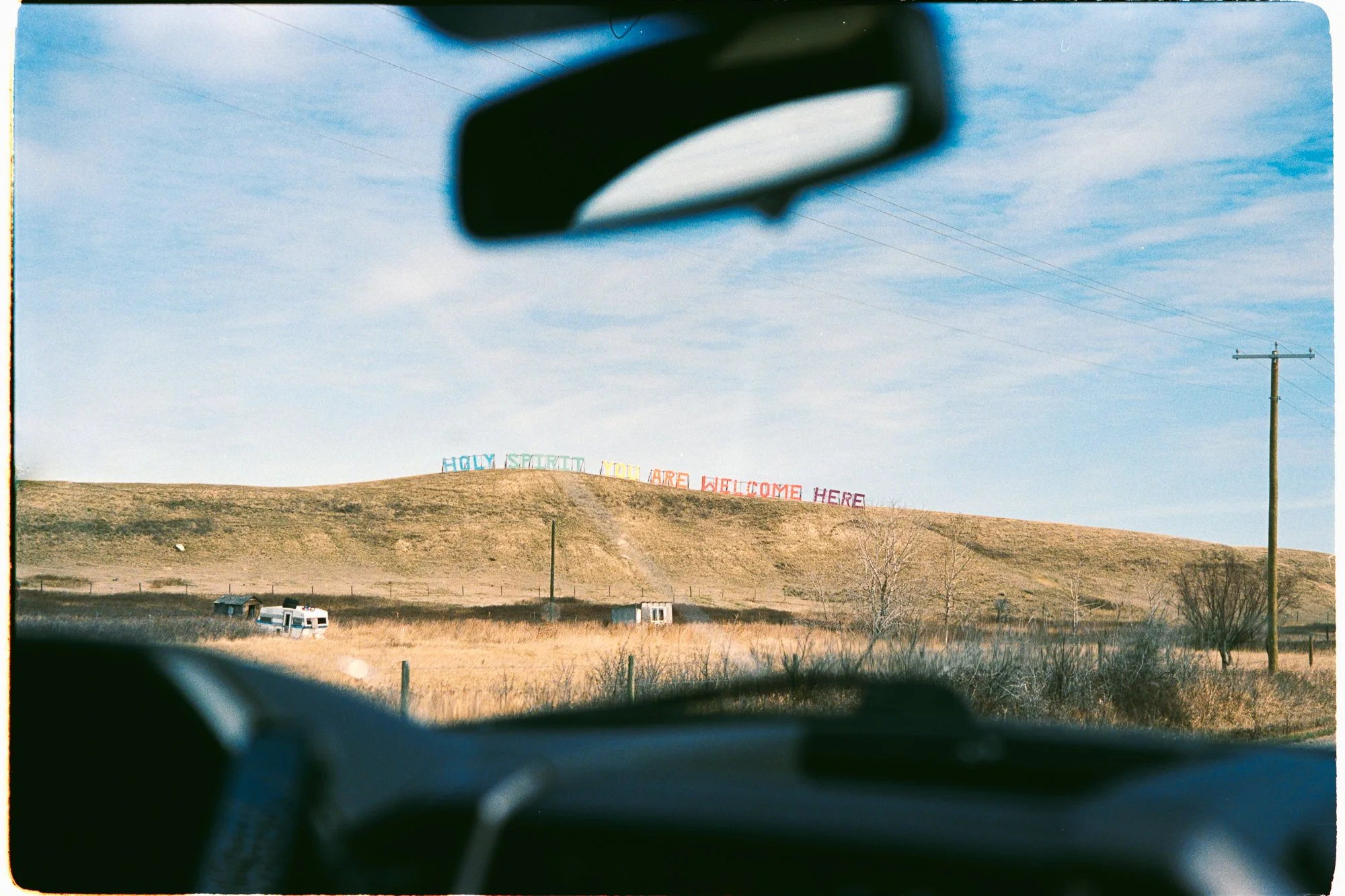 Prairie landscape photographed through the windsheild of a truck.  A rainbow sign sits large on the horizon saying 'Holy Spirit you are welcome here'.  Two camping trailers and power lines stand in the foreground.  Photographed on Kodak Gold 35mm.