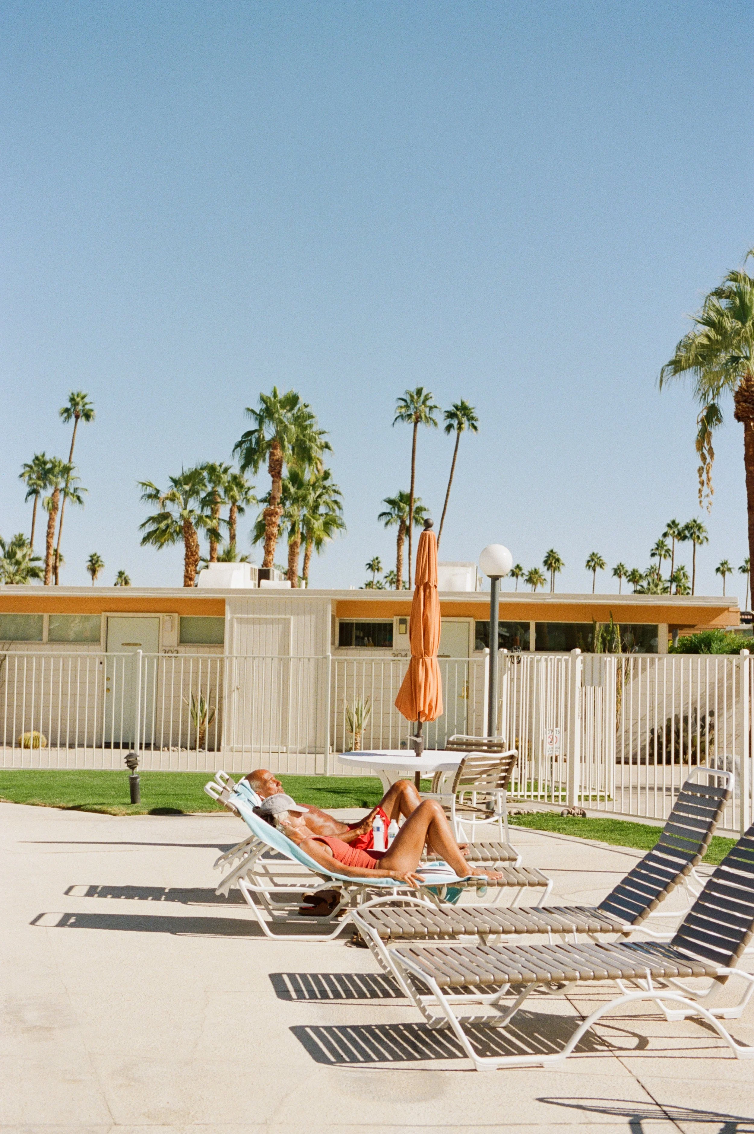 Two sunbathers relax on loungers in front of mid-century apartments and a champagne cork pool at ocotillo lodge in Palm Springs, California.  Photographed on 35mm film.
