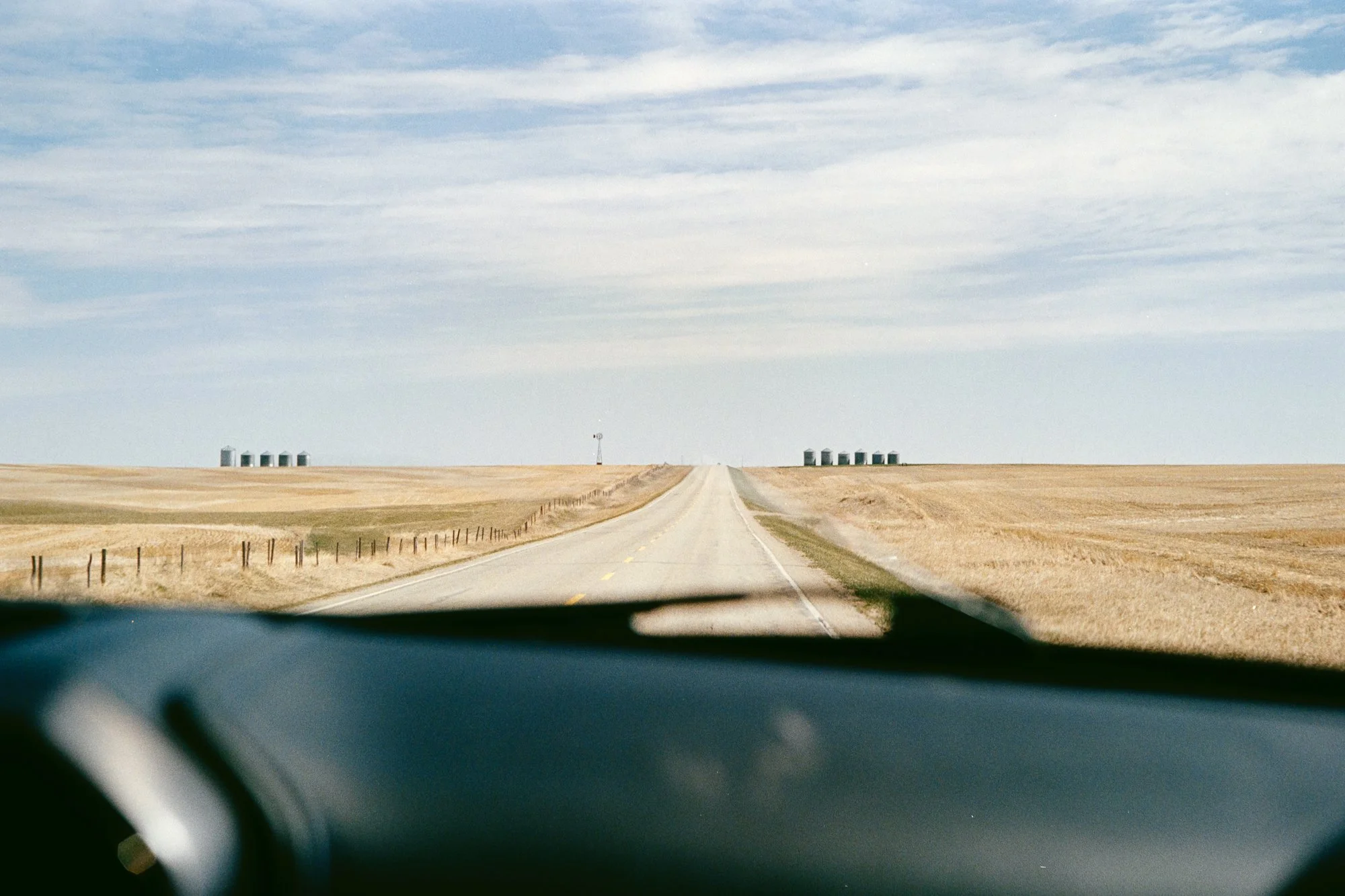 Colour film photograph of two sets of silos on the horizon, divided by a highway.  Photographed through the windsheild of a truck in Southern Alberta.  Photographed on Kodak Gold 35mm film and developed at home.