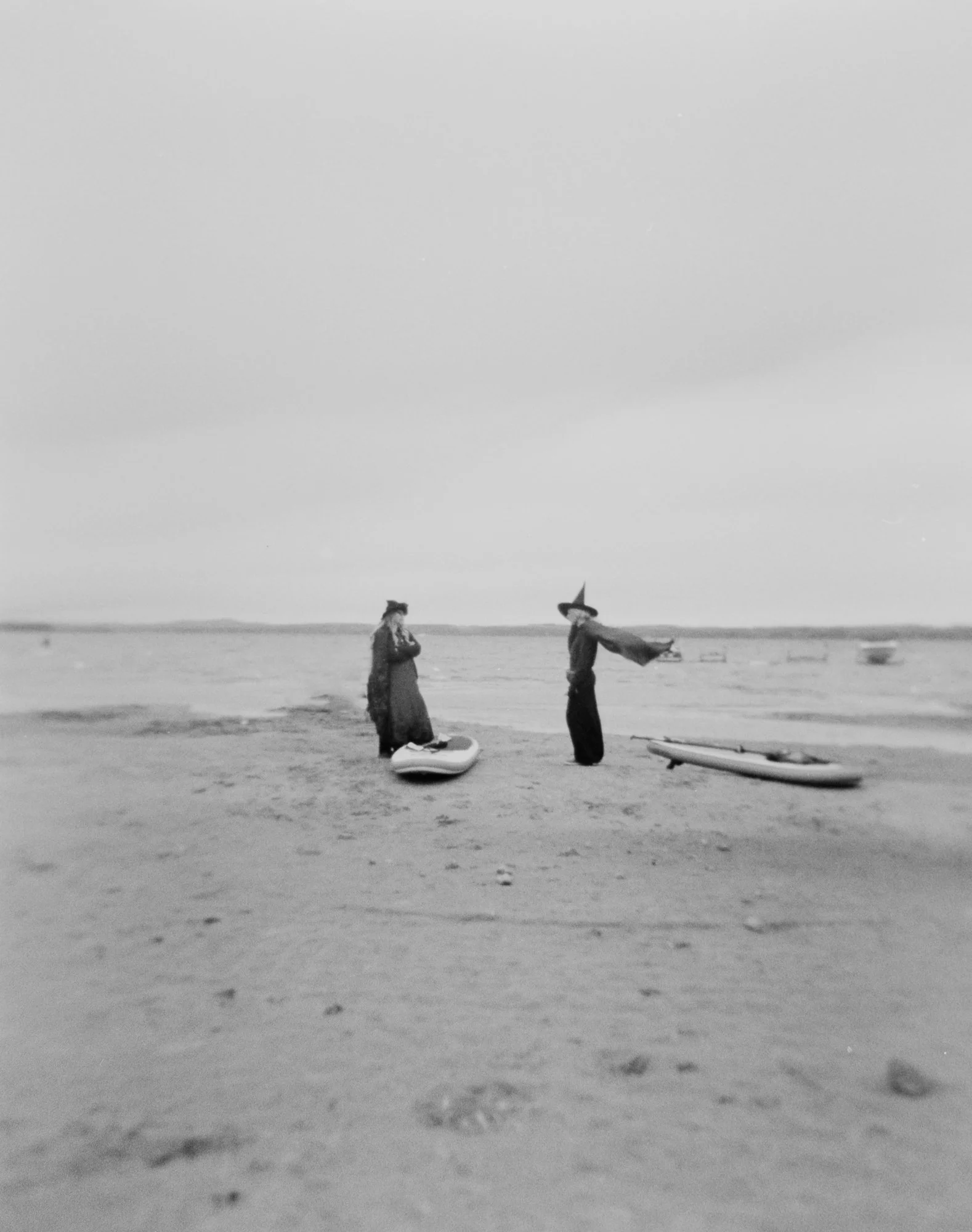 Two women dressed as witches stand on the beach with paddleboards. One witch's cloak is flying behind her caught in the wind.  Photographed on 120 film with a Holga Camera.