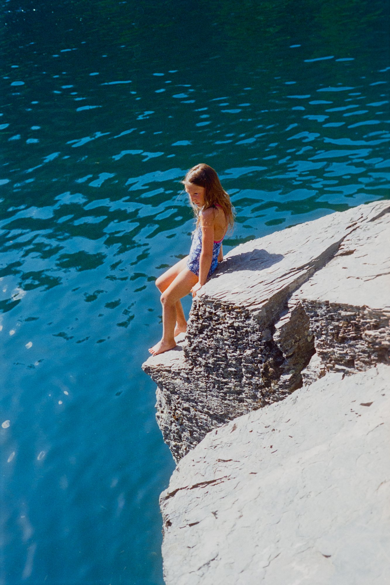 A girl in a vlue swimsuit stands on the edge of a cliff , readying herself to jump into vibrant blue waters.  Photographed on 35mm film.