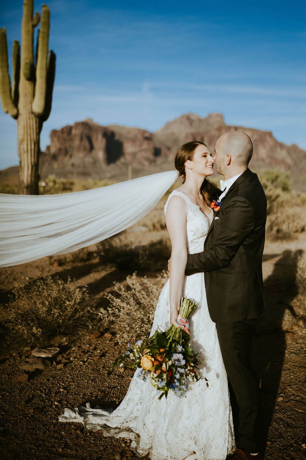 Bride and groom kissing in the desert with cactus and mountains in the background