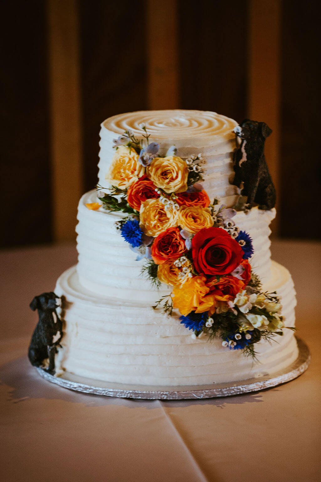 Wedding cake with vibrant flowers and two dog figurines that look like they're eating the cake