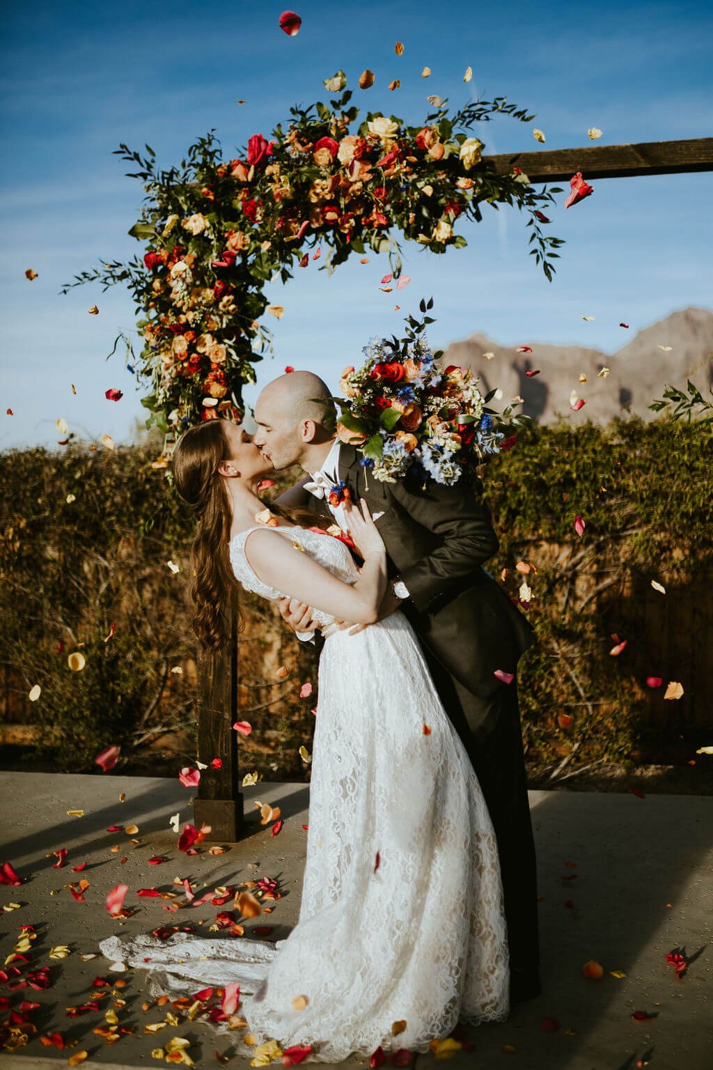 Bride and groom kissing under arbor with flower petals in the air and mountains in the background at Arizona desert wedding