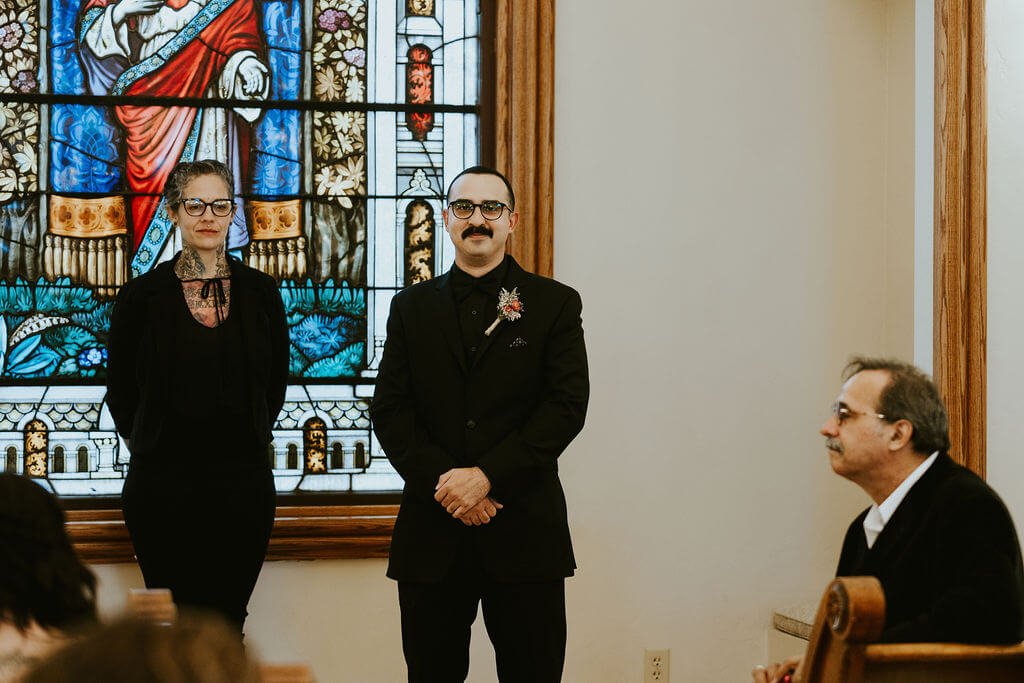 Groom and wedding officiant standing at the front of a chapel with beautiful stained glass in the background