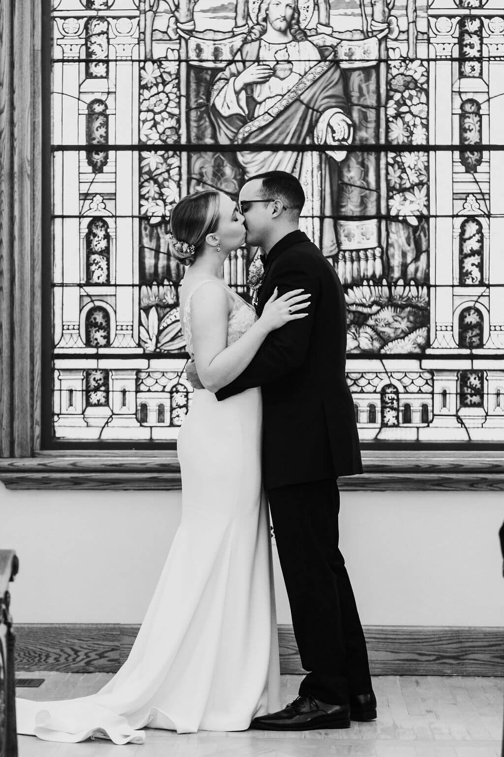 Black and white photo of bride and groom kissing in front of stained glass in a chapel