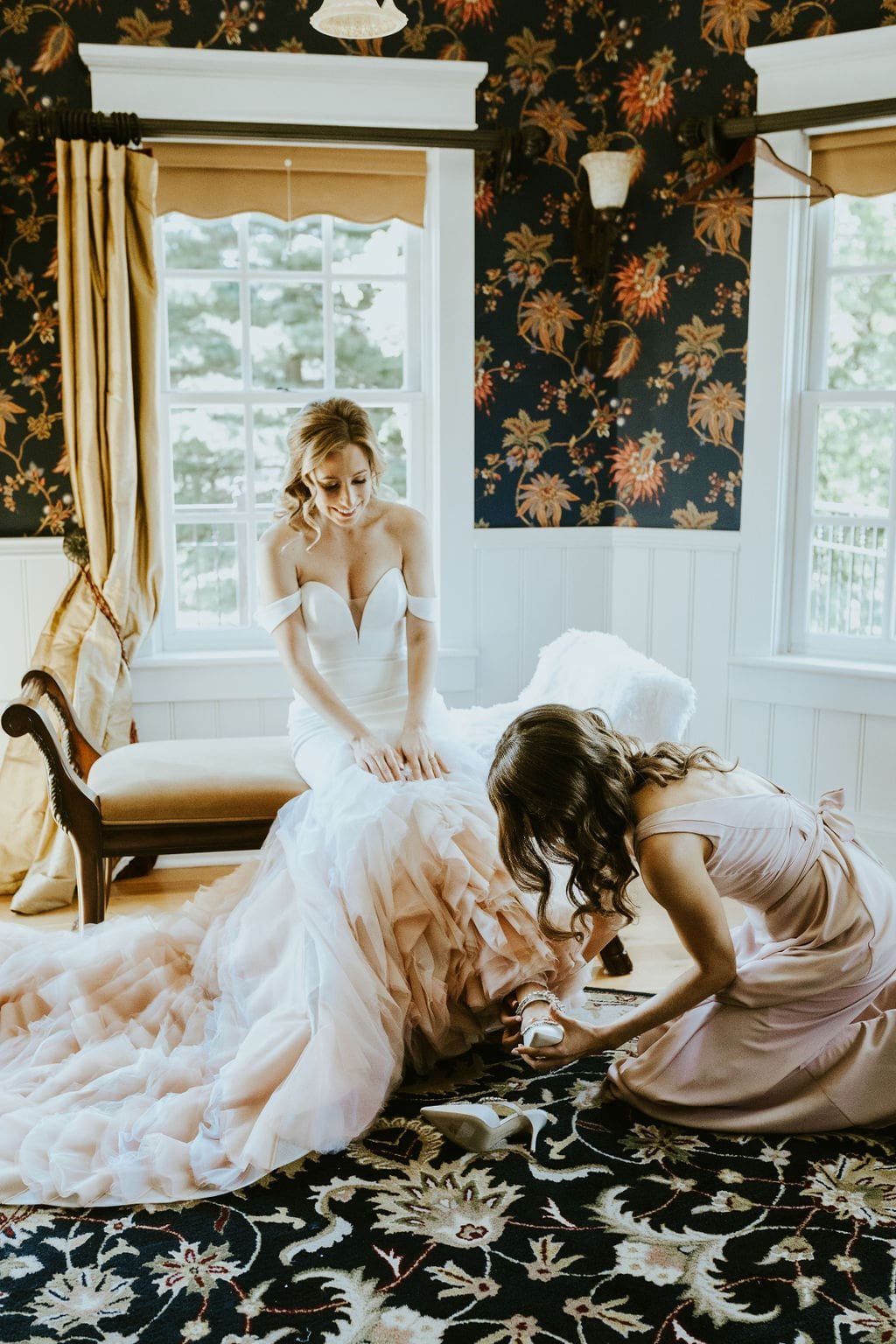 Bridesmaid helping put shoes on bride while getting ready for classic wedding