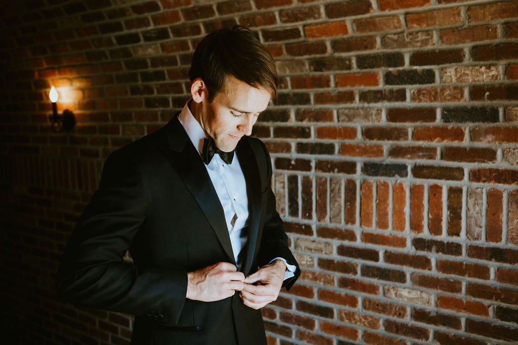 Groomsman buttoning black suit while getting ready for classic farmhouse wedding in Illinois