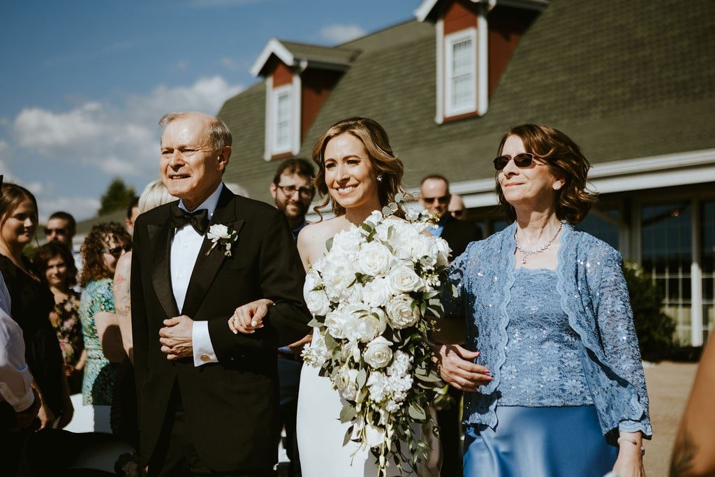 Bride walks down the aisle with her parents at an outdoor wedding ceremony at Orchard Ridge Farms