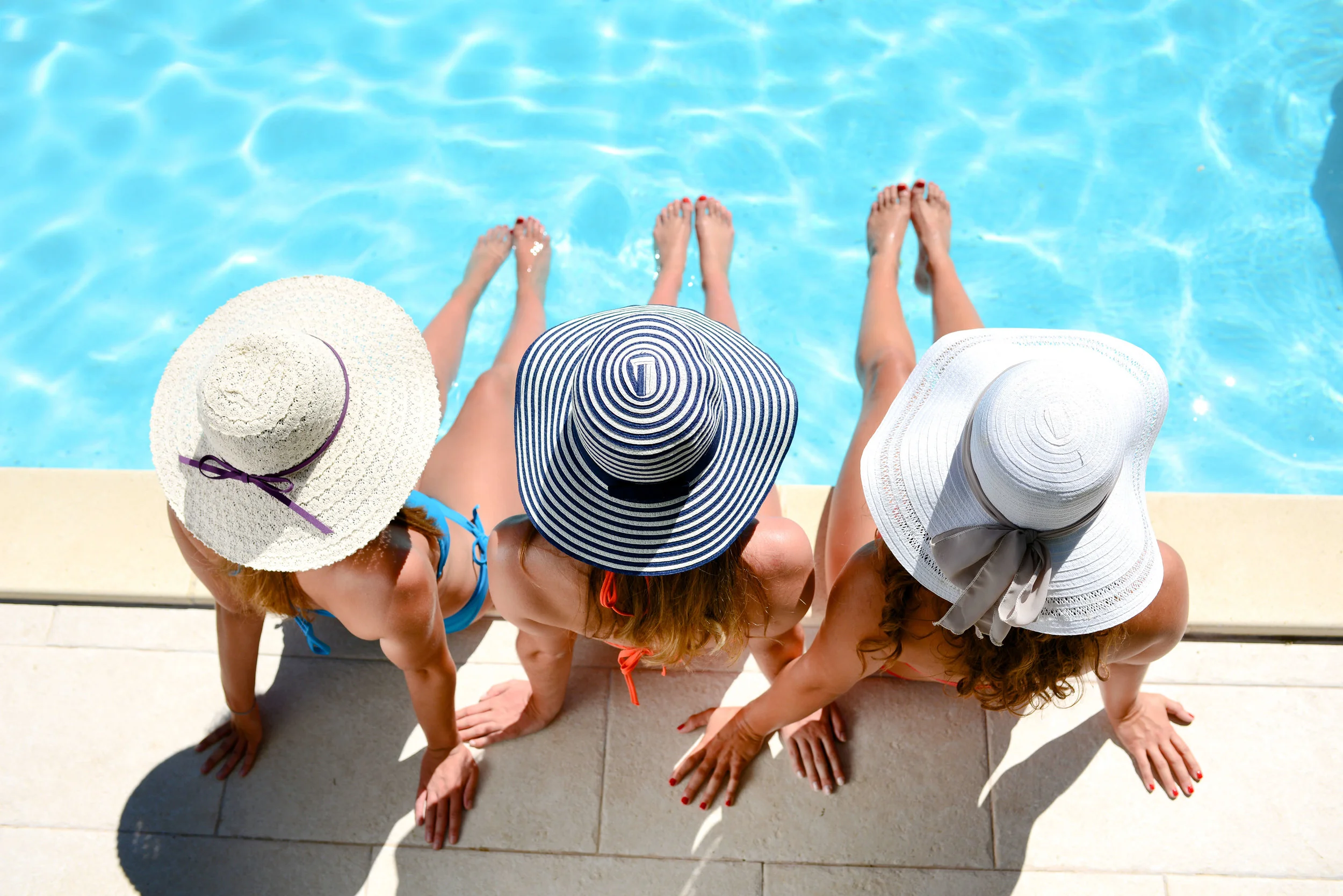 Three women wearing sun hats sitting by a well-maintained swimming pool with their feet dipped in the water.