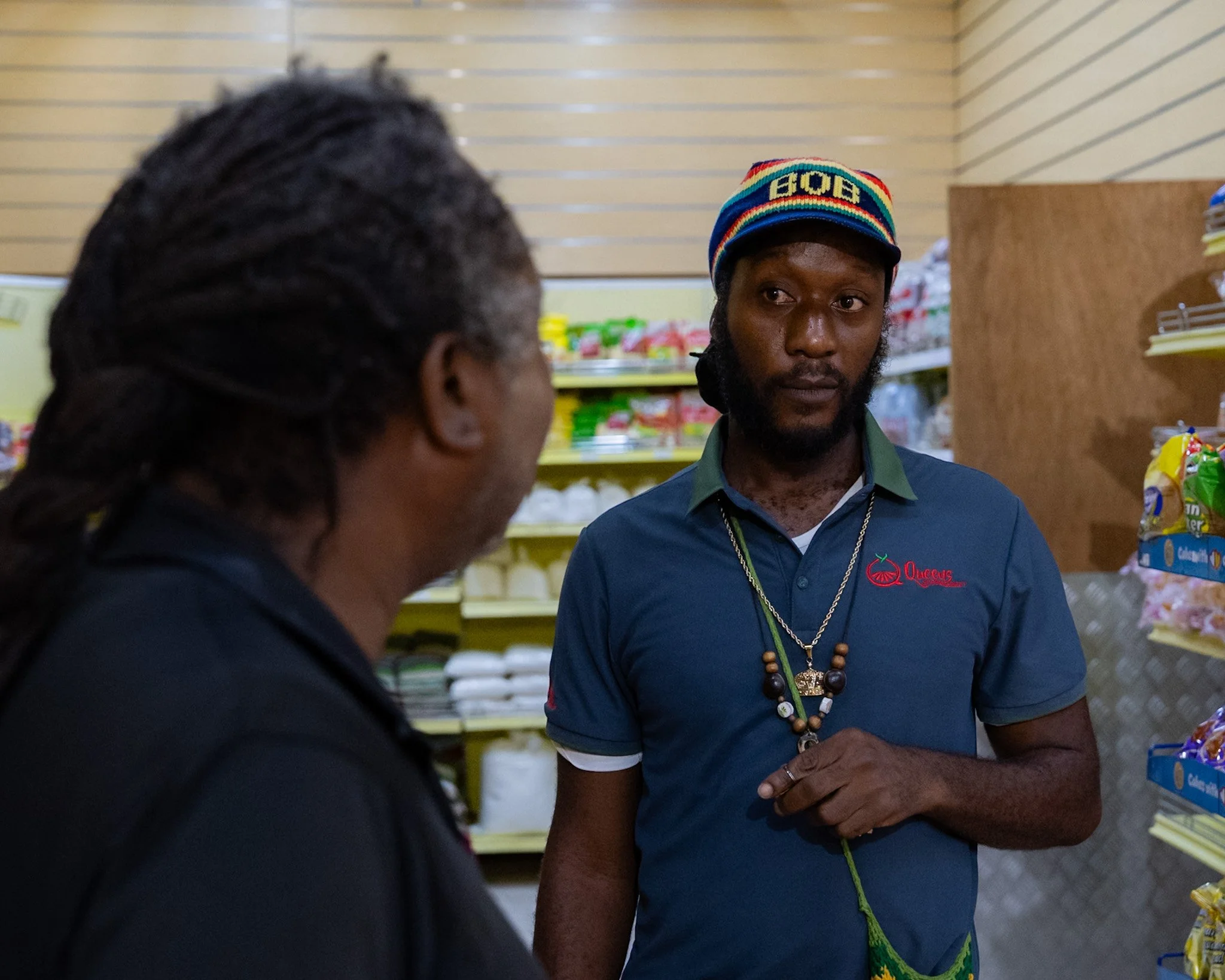 Daniel “Ras Daniel” Smith-Cooke, 2nd-generation Rastafari assistant manager, speaking with a customer inside a Jamaican supermarket.