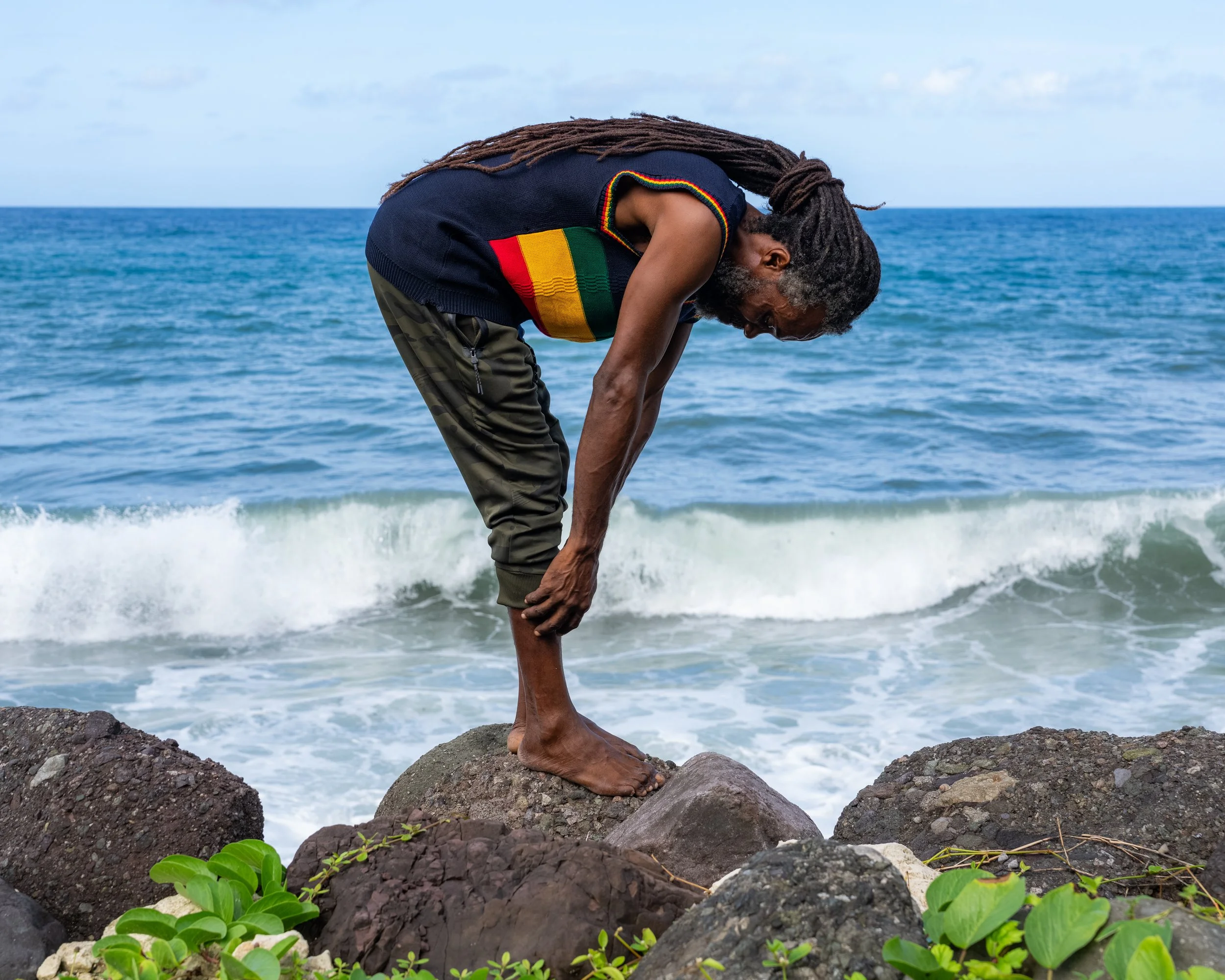 Joel “Ras Binghi Pele” Dyke, 3rd-generation Rastafari, bending over rocks by the sea in Jamaica with dreadlocks falling forward.