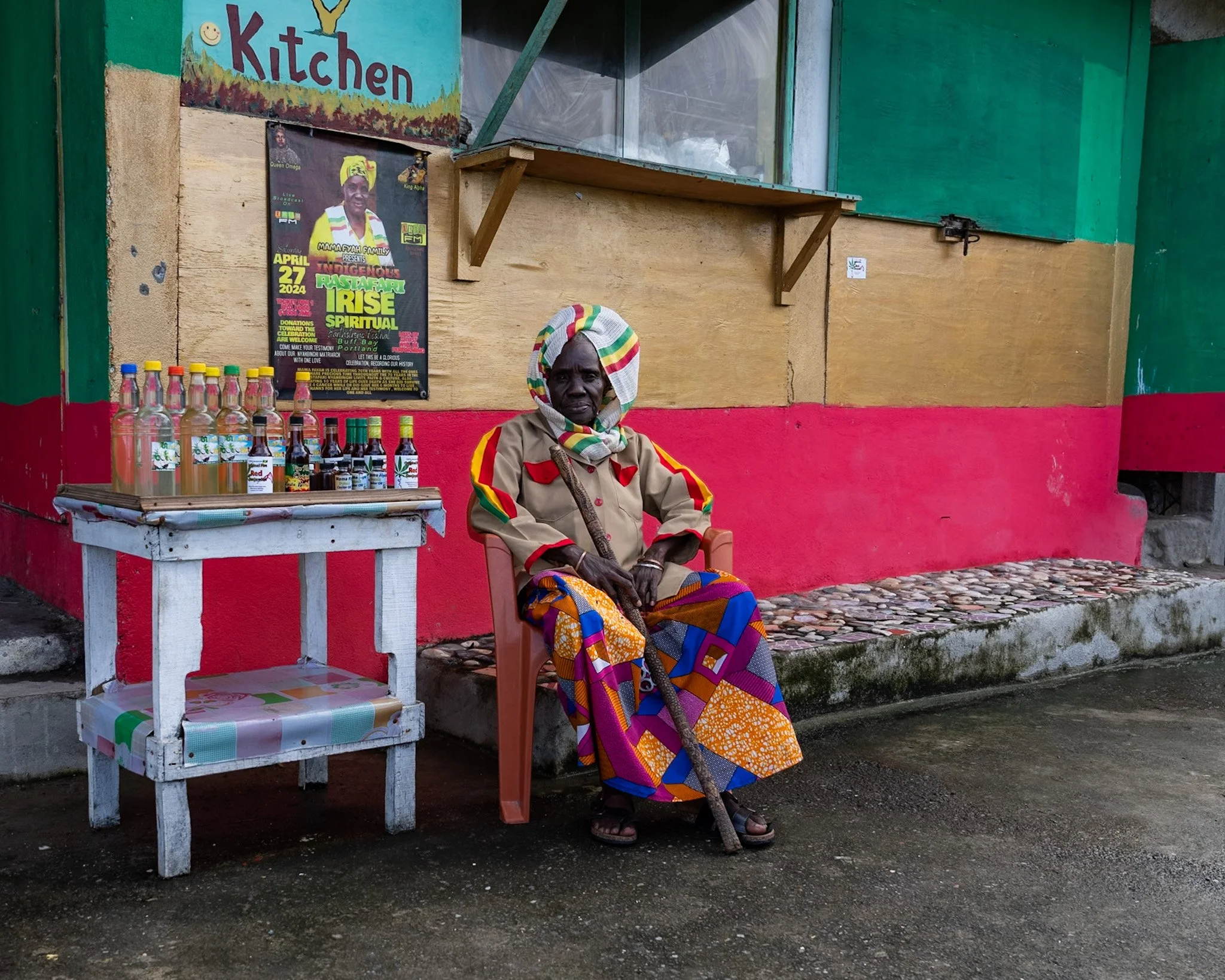 Pearline “Mama Fiyah” Wolfe, 2nd-generation Rastafari spiritual leader, seated outside a colorful building in Jamaica wrapped in patterned fabric.