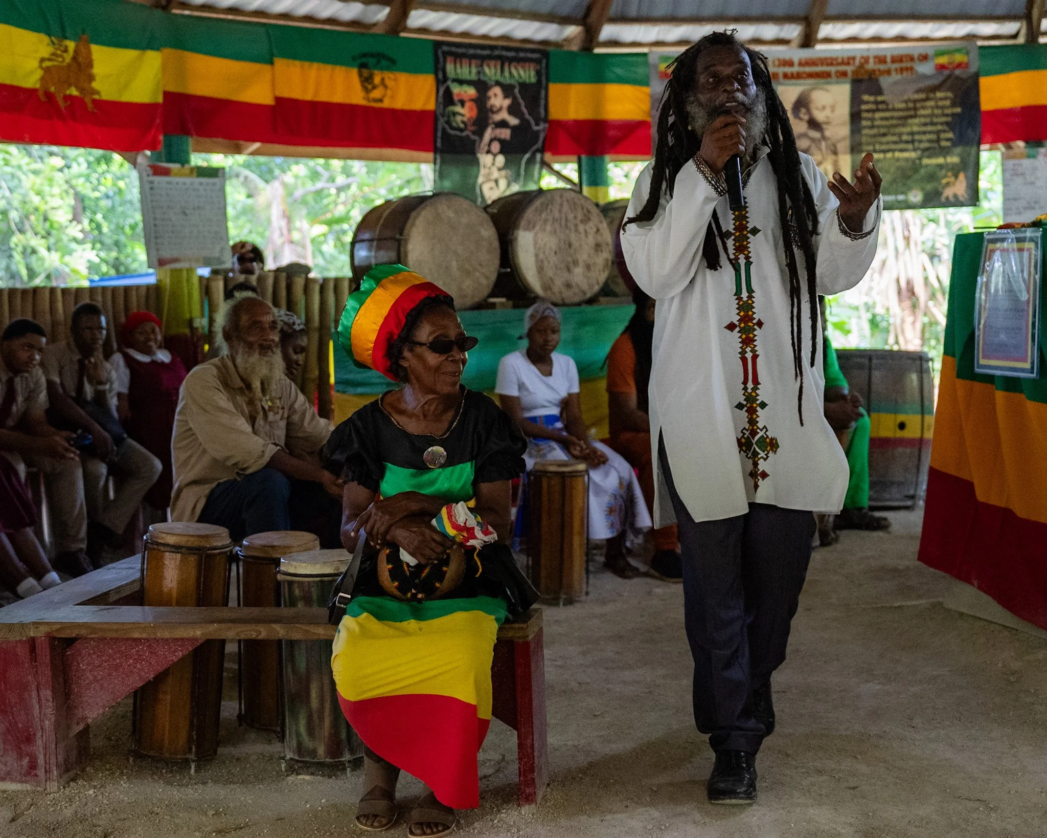 Donavan “Ras IVI” Wright, Rastafari high priest and advocate, speaking inside a Nyabinghi space with drums and Rastafari flags in Jamaica.