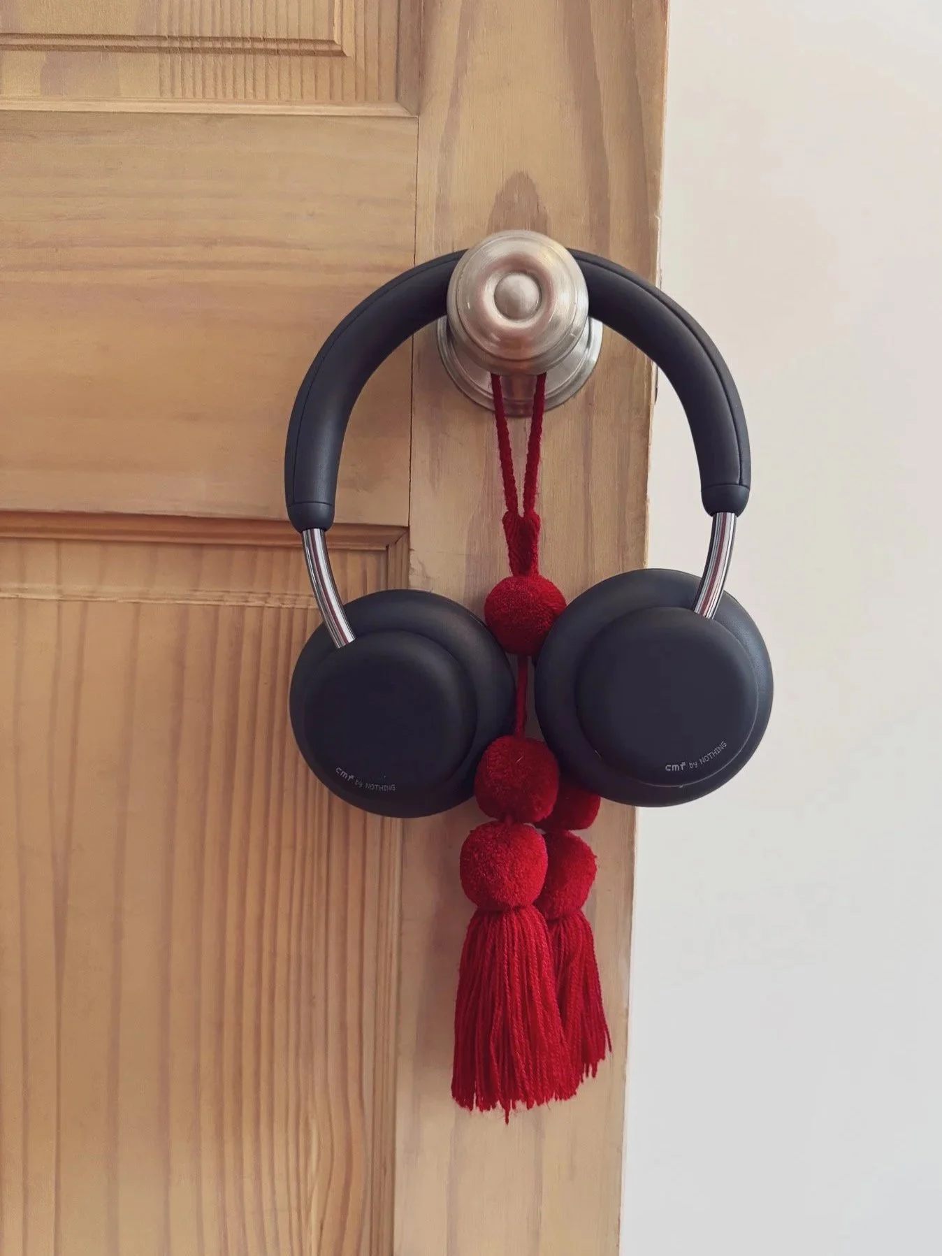 A pair of black over-ear headphones hanging on a wall hook, decorated with a red tassel and pom-poms.