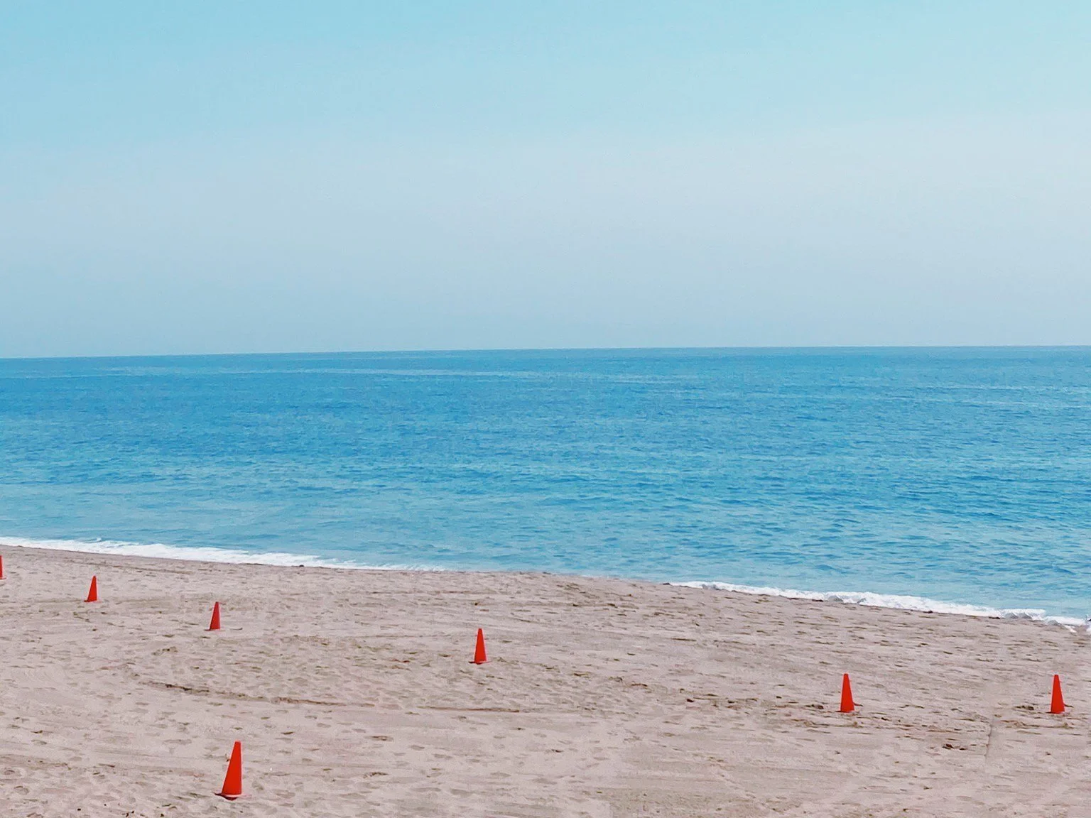Empty sandy beach with orange cones and a calm blue ocean under a light blue sky.