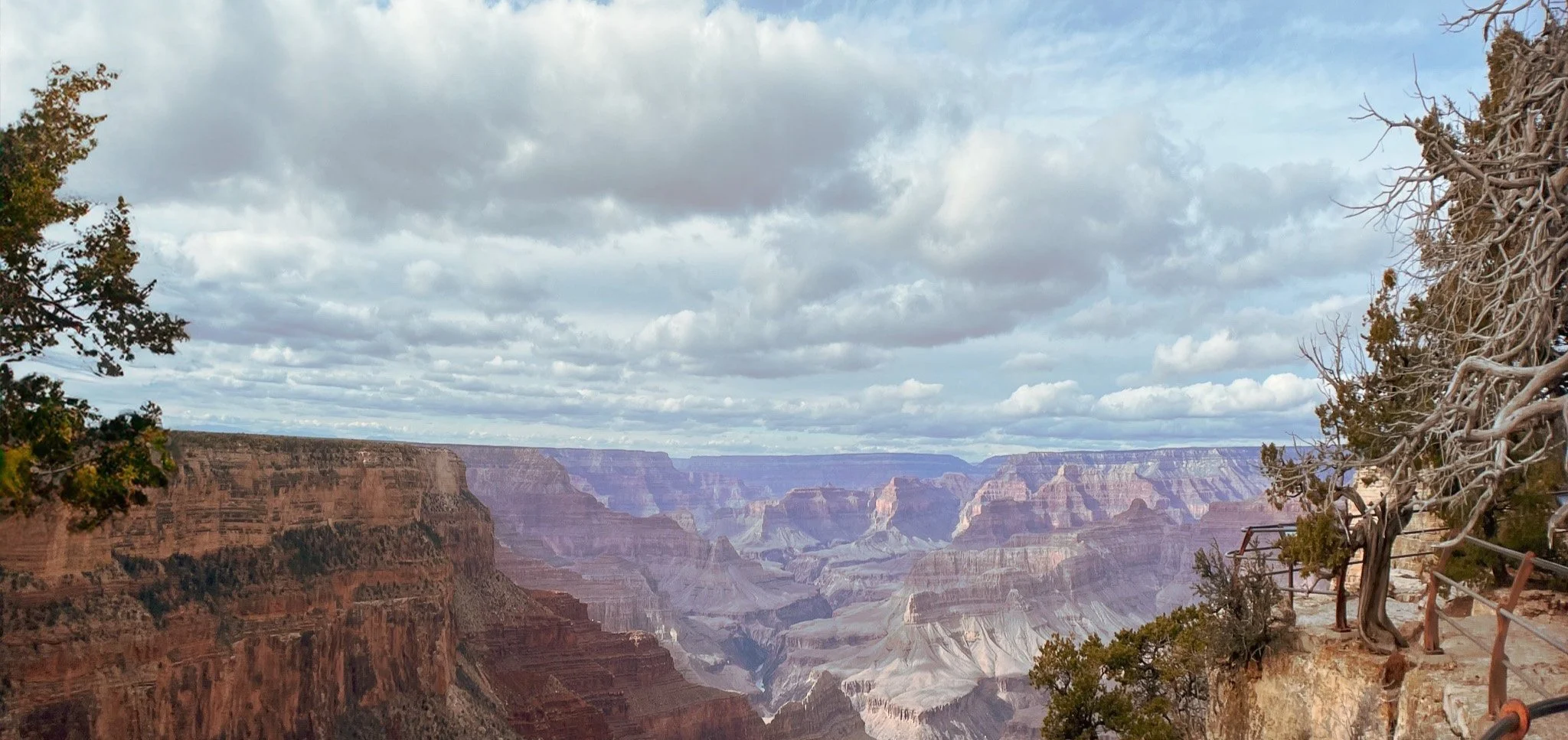 View of the Grand Canyon with layered red rock formations, cloudy sky, and a pathway with a railing on the right side