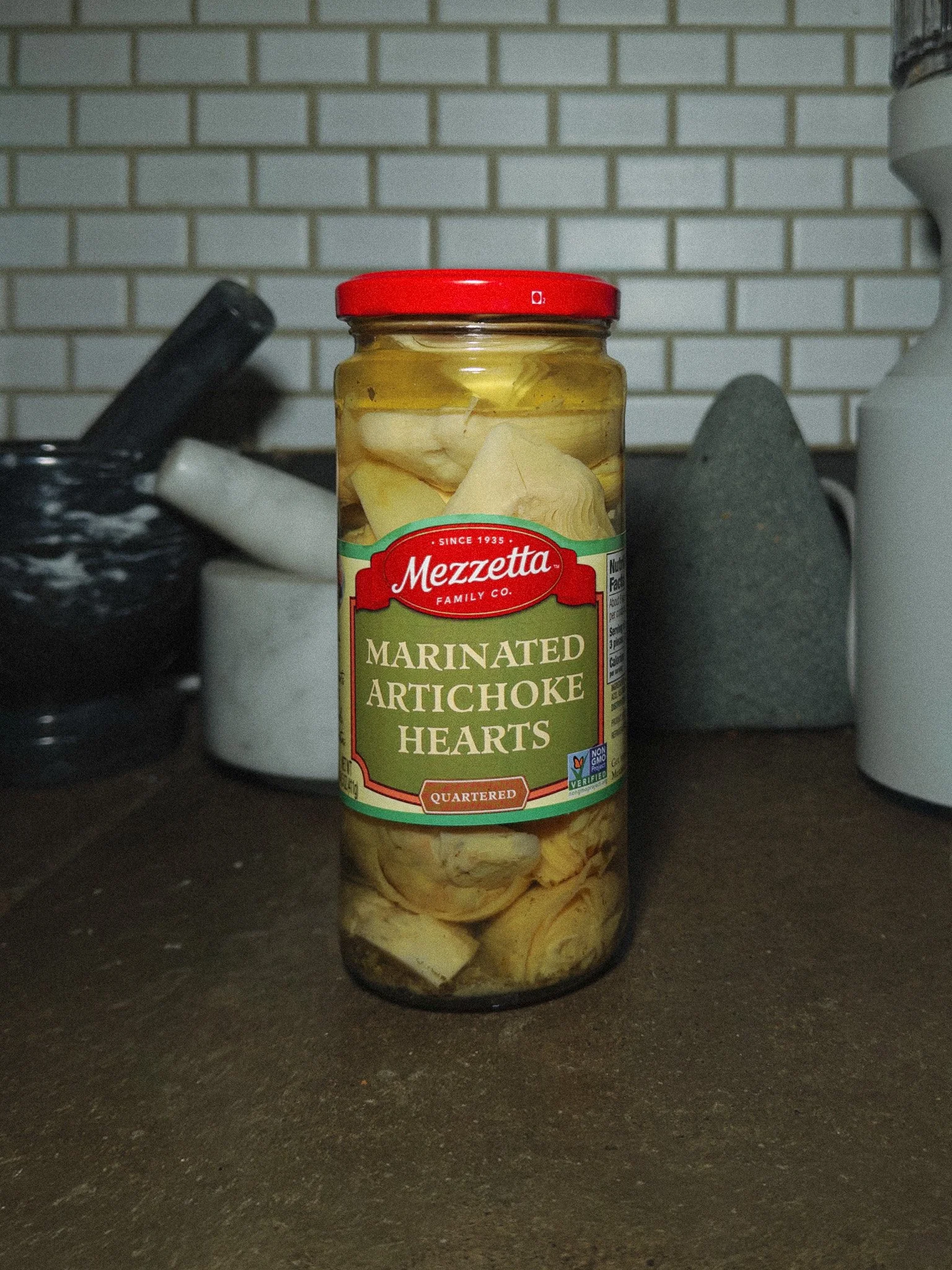 A jar of Mezzetta marinated artichoke hearts on a dark kitchen countertop, with a white brick wall in the background.
