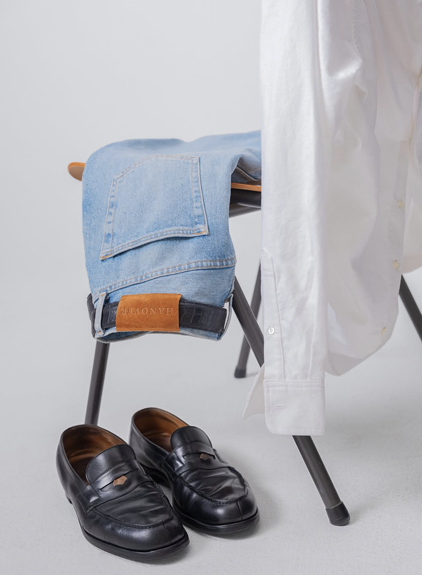 A pair of black loafers on a light gray floor, a white shirt hanging from an unseen chair, and a folded pair of light blue jeans with a leather patch labeled 'HOUN' hanging over a black chair.