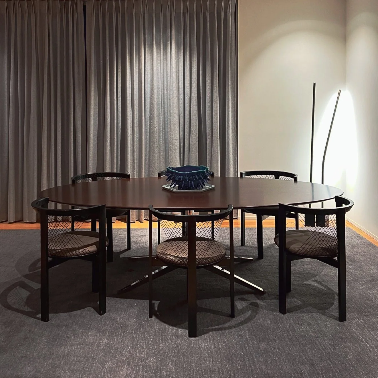 Modern dining room with a dark oval table, six mesh-backed chairs, a decorative centerpiece, and a floor lamp, set against heavy curtains.