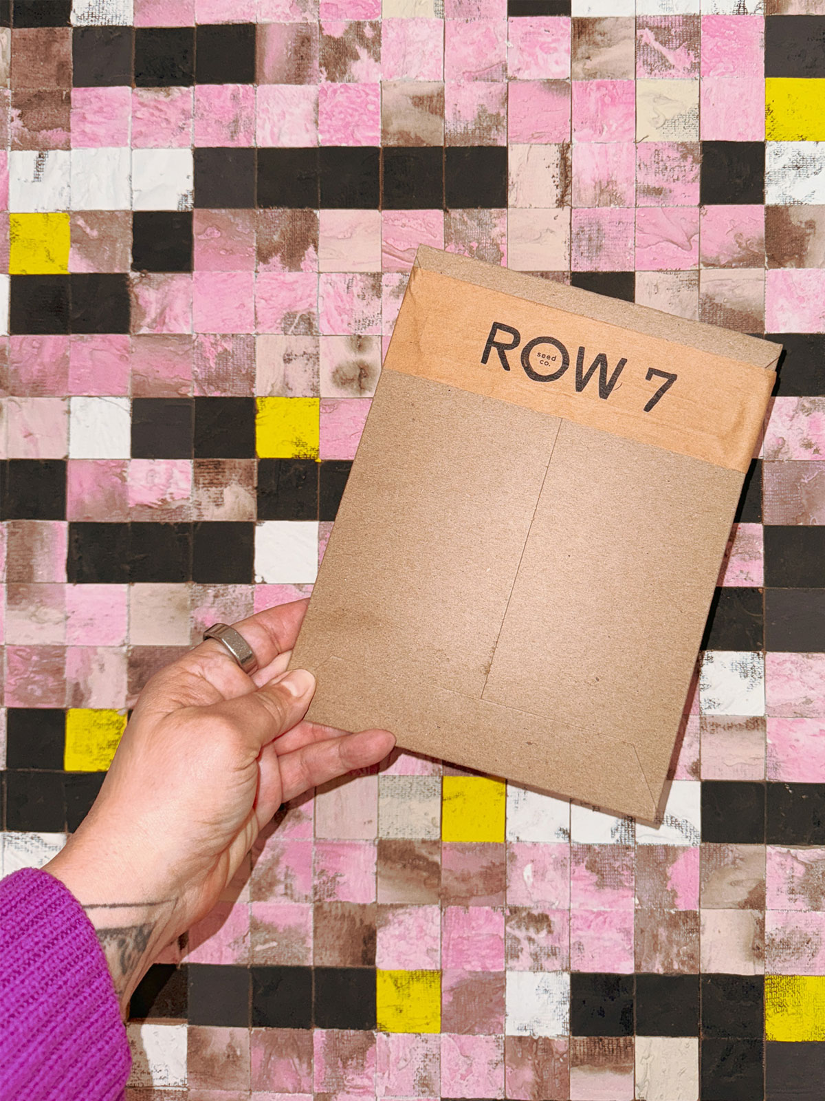 Hand holding a brown paper plant tag labeled 'ROW 7' in front of a colorful tiled wall.