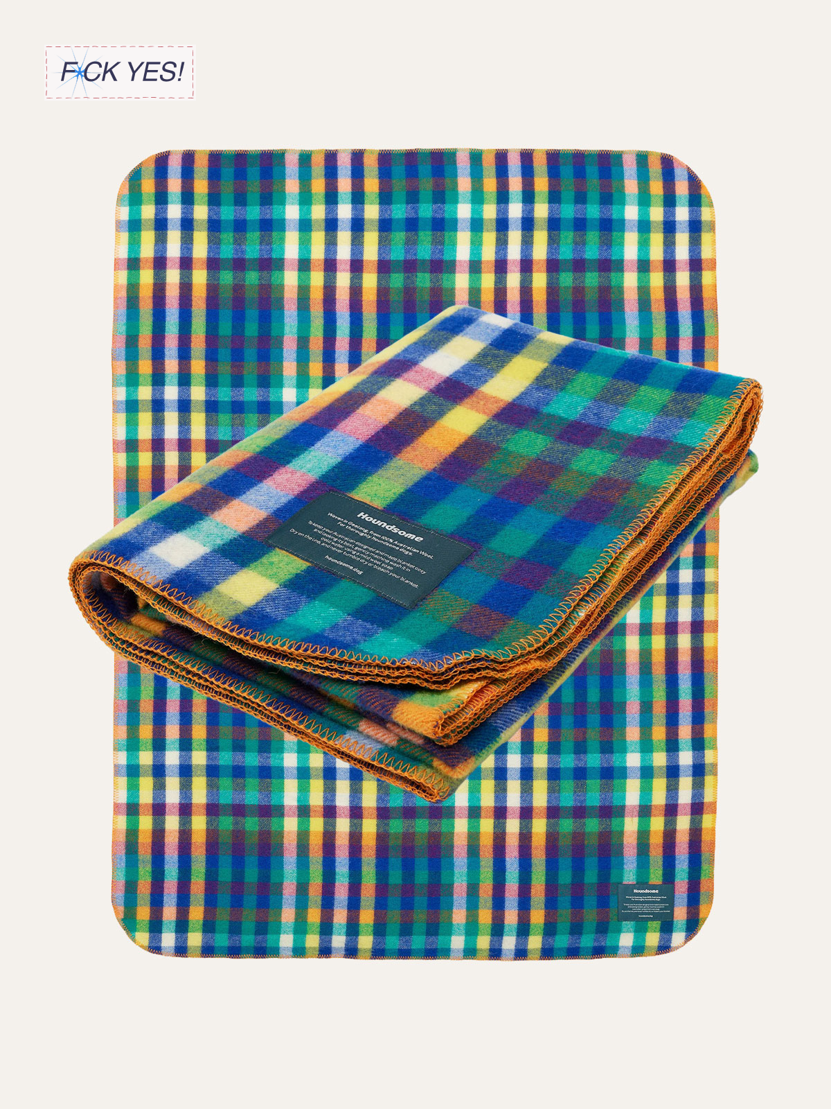 Colorful plaid blanket with a black label over a matching plaid blanket on a chair with a matching plaid cushion in the background. Text in the top left corner reads 'F*CK YES!'.