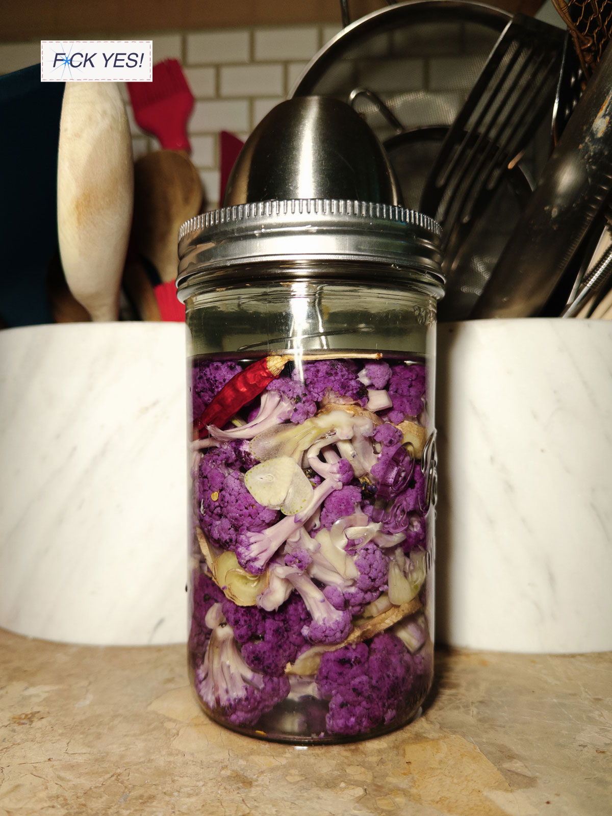 A jar filled with purple cauliflower florets, sliced garlic, and red chili pepper, placed on a kitchen counter with kitchen utensils in the background.