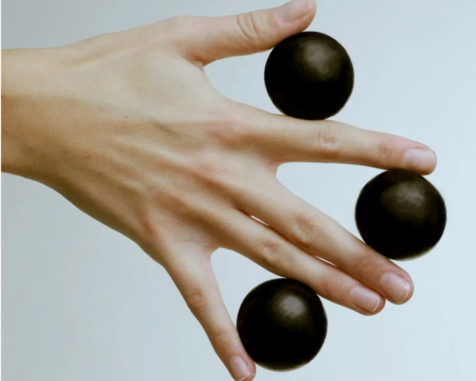 A hand with three black spherical objects balanced on different fingers against a plain light background.