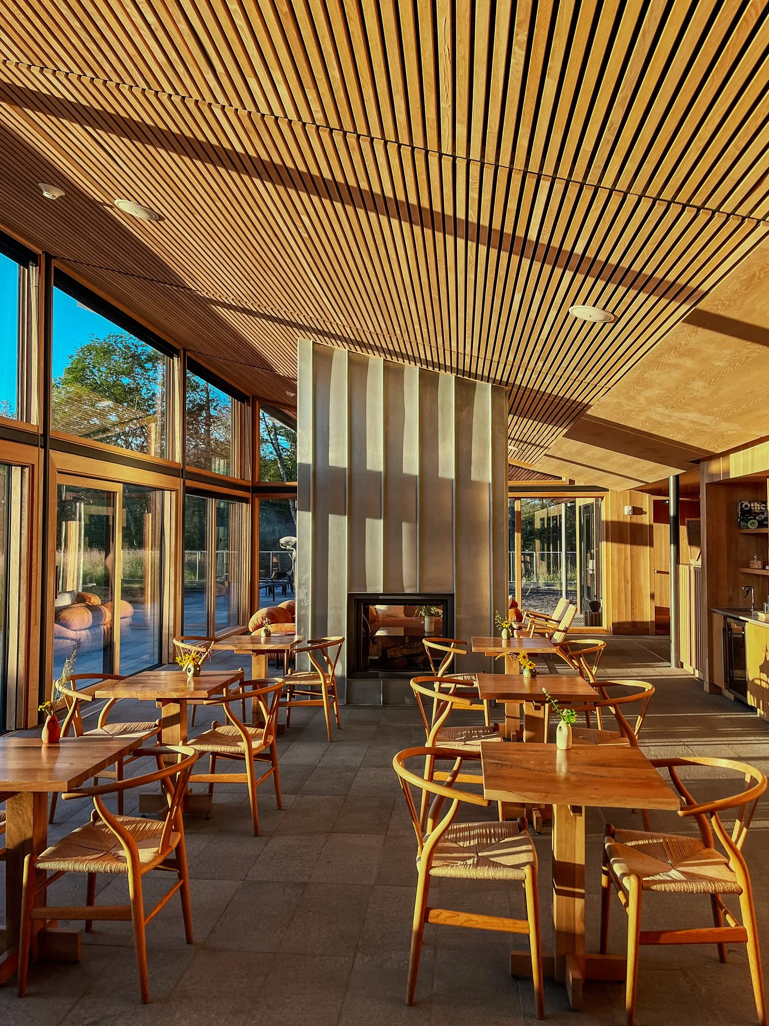 Interior of a modern restaurant with wooden tables and chairs, large windows, a fireplace, and a wood-paneled ceiling.
