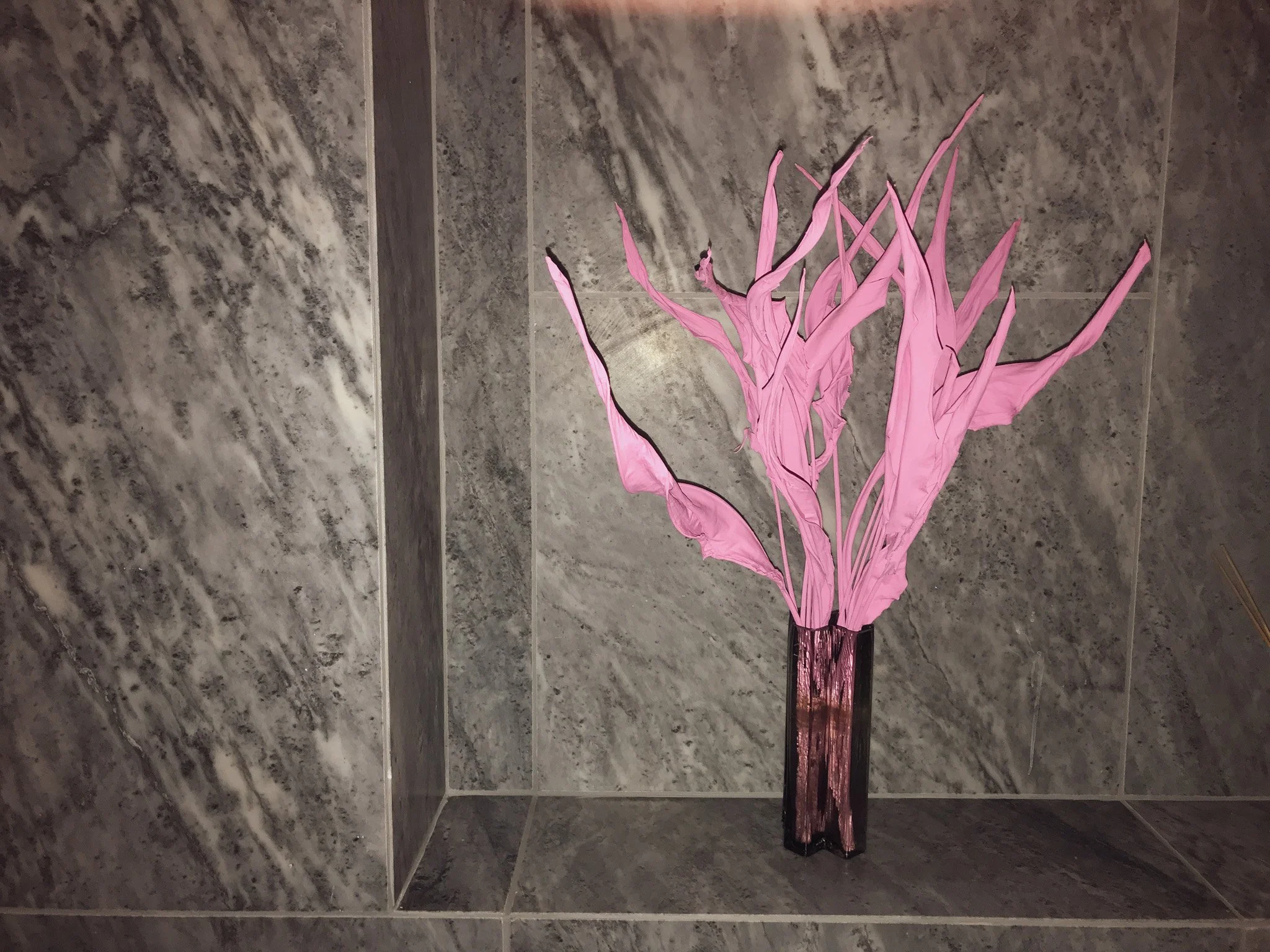 A decorative pink paper or fabric flower arrangement in a tall black vase placed on a gray marbled tiled surface.