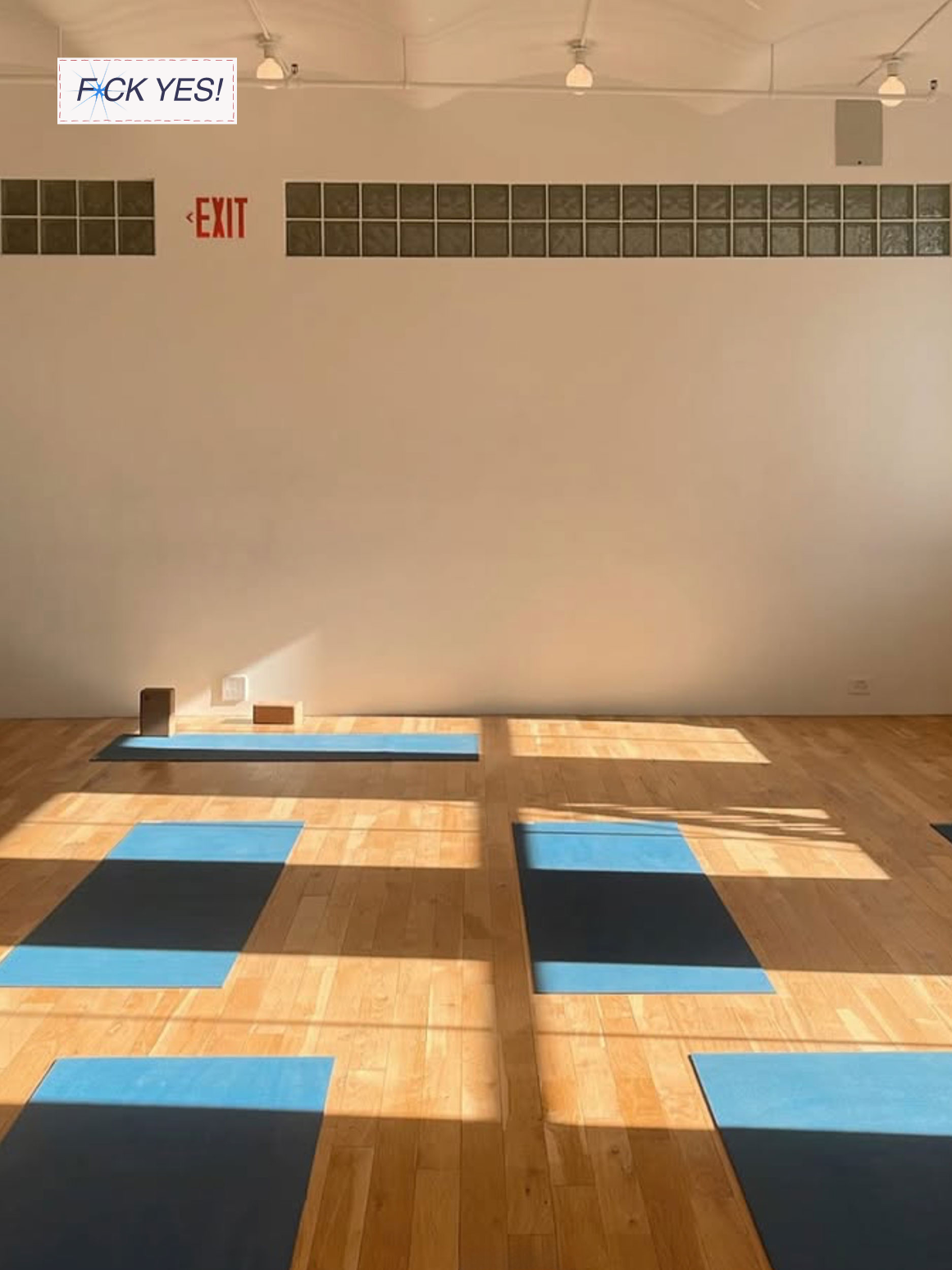 Yoga mats and yoga blocks on a wooden floor in a studio with sunlight casting shadows, and a large blank wall with an exit sign and a motivational sticker that says 'F*** YES!'