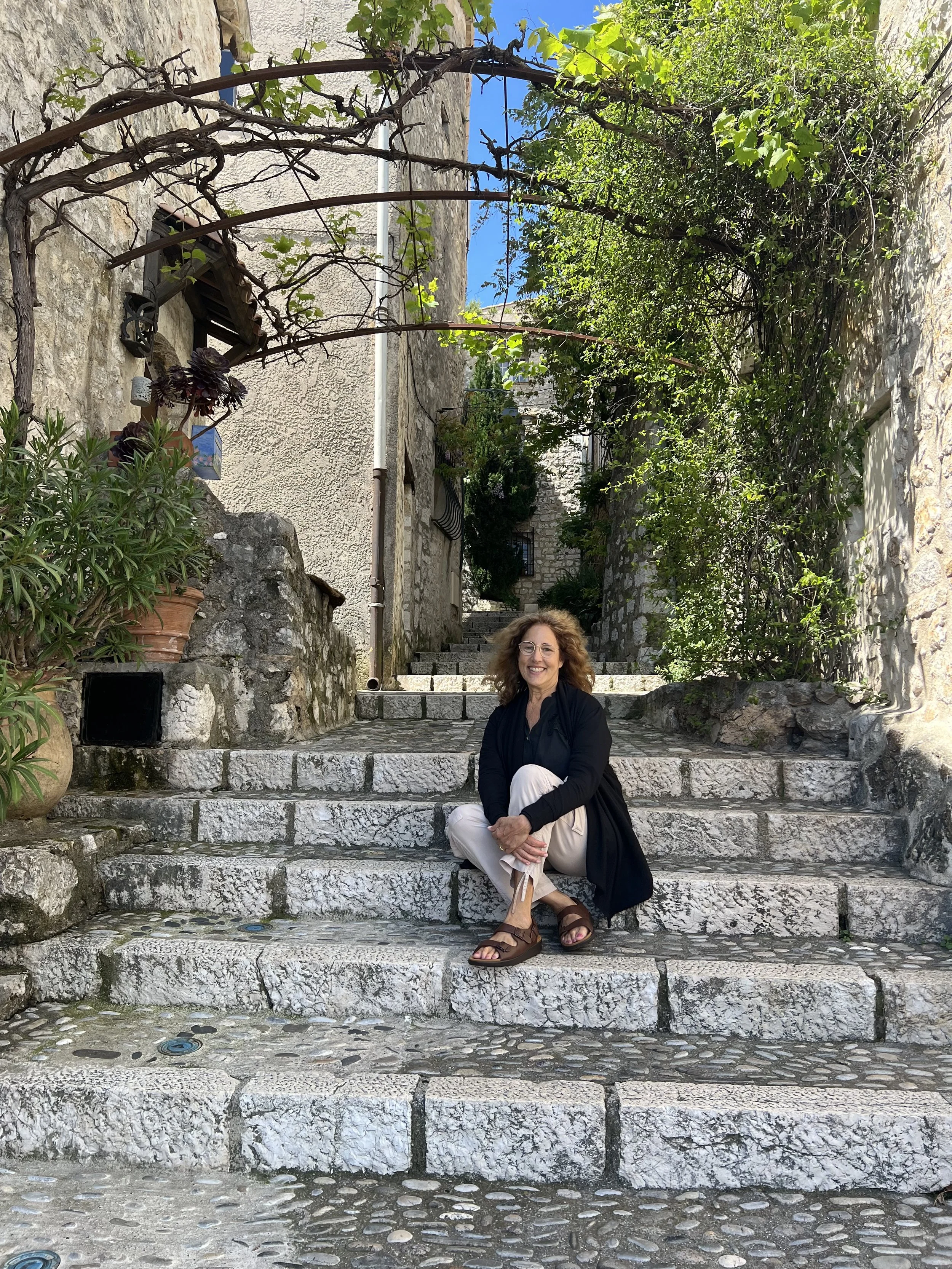 Woman with brown curly hair and glasses sitting on stone stairs in a narrow outdoor alleyway with stone walls, greenery, and blue sky overhead.