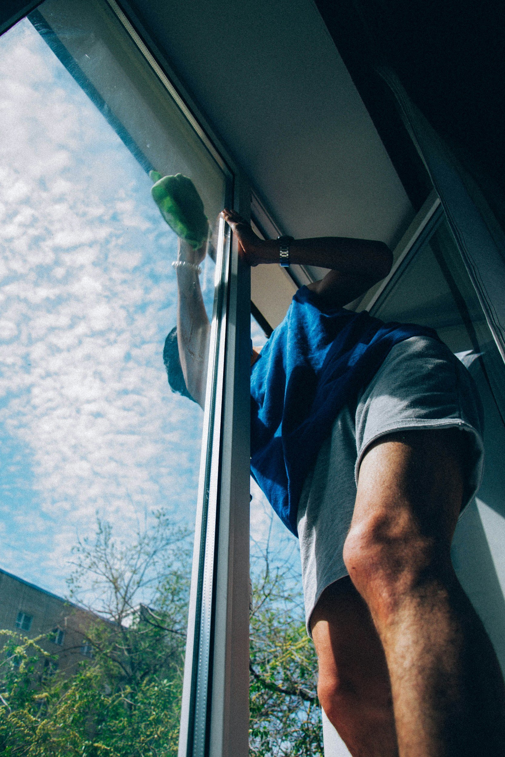 Man standing by an open window cleaning the glass with a cloth, with blue sky and trees visible outside.