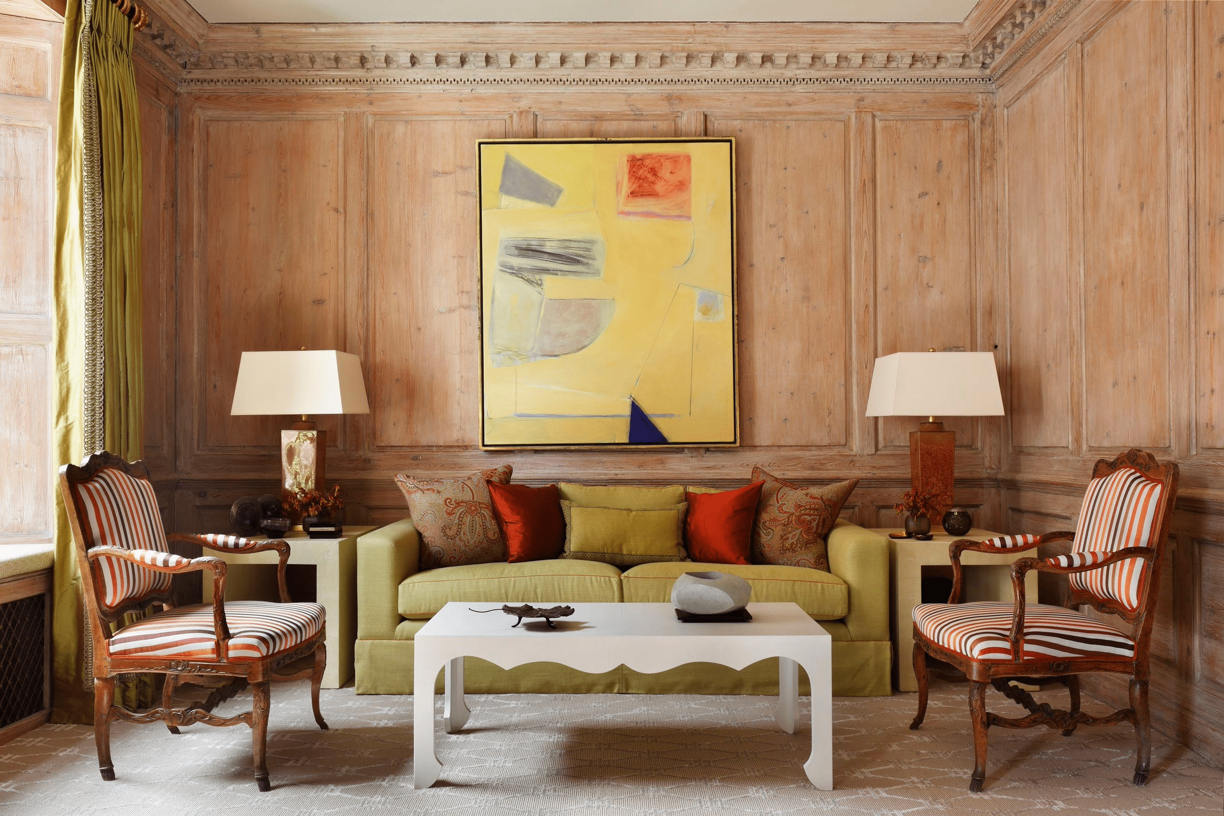 A cozy living room with a green sofa, striped armchairs, abstract painting, and wood paneling, blending modern and traditional styles. Designer: Mark Gillette. Photography by Alexander James.