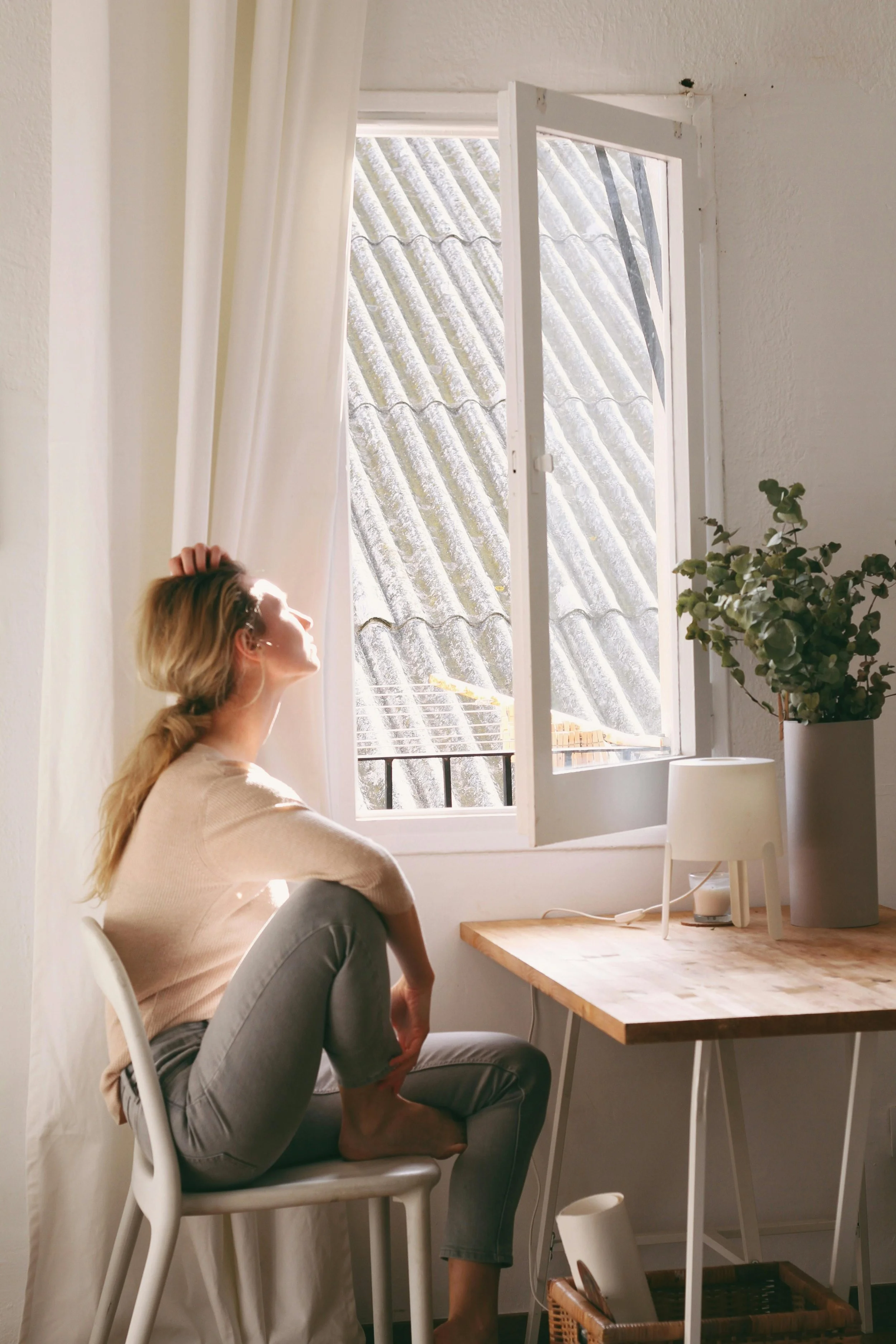 Woman sitting in a chair angled toward an open window with sunlight streaming into the room.
