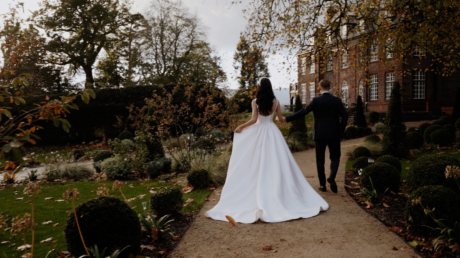 Bride and groom walking hand in hand on a garden path near a large brick building with trees and bushes.