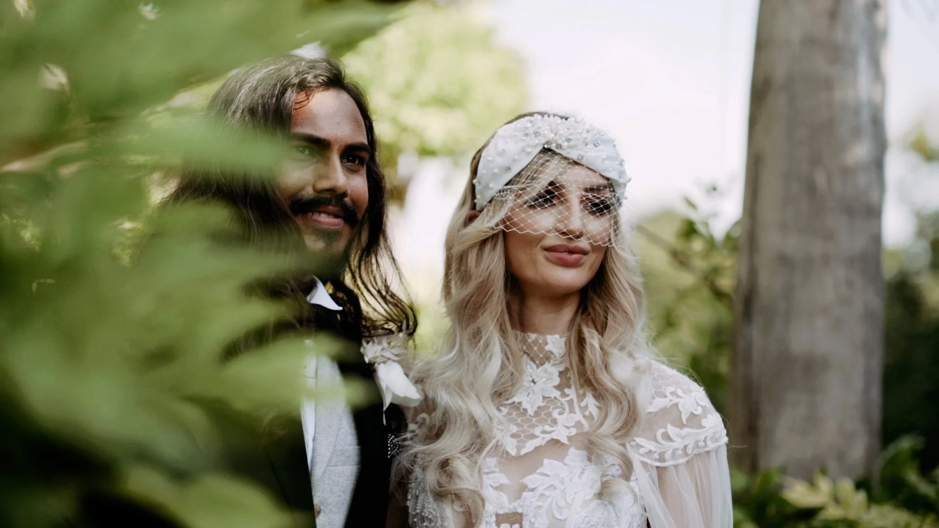 A couple dressed in wedding attire standing outdoors with greenery, trees, and sunlight in the background.