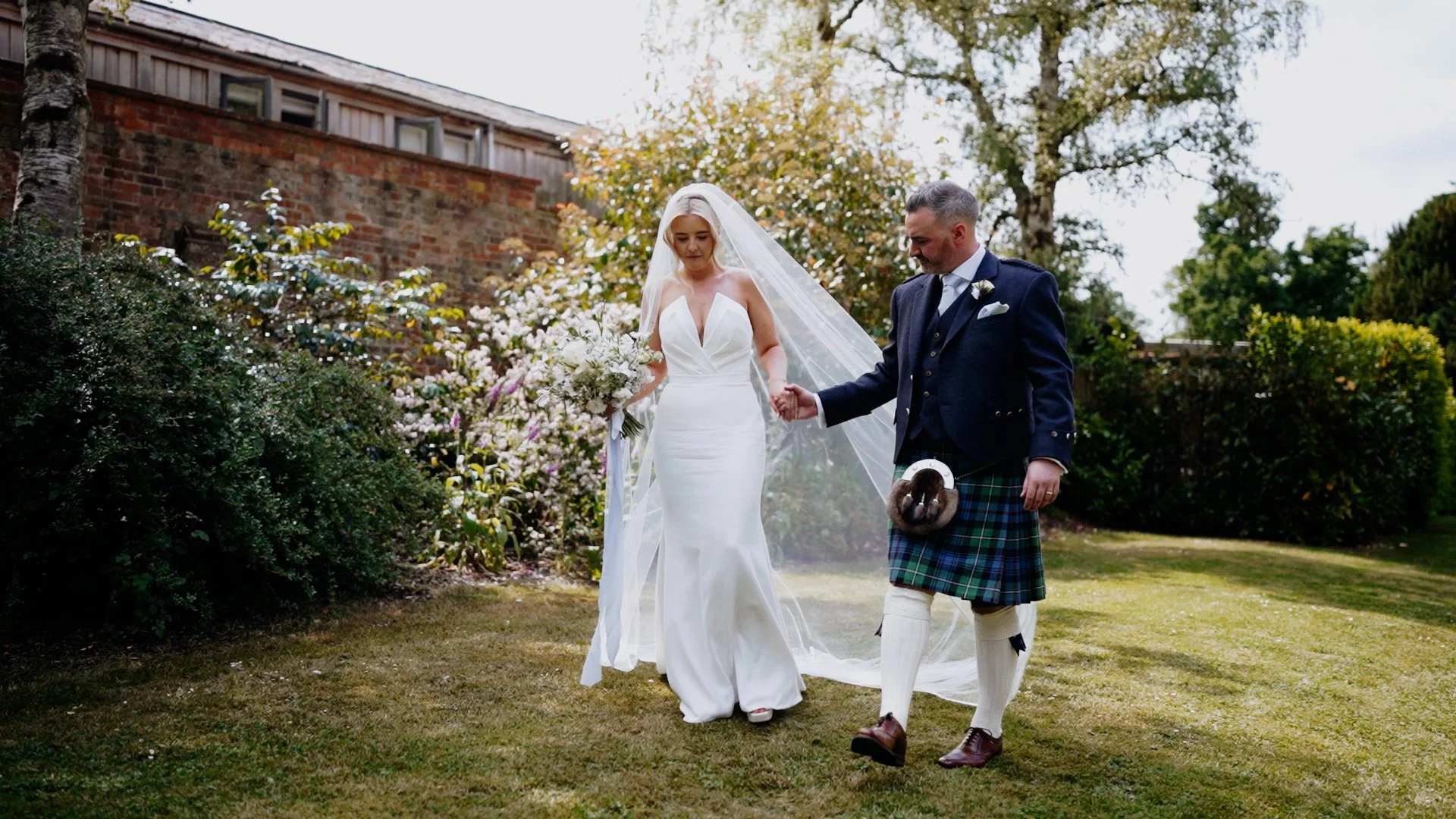 Bride in a white wedding dress holding a bouquet standing next to a groom in a traditional kilt and jacket, who is holding her hand in a garden with trees and bushes.