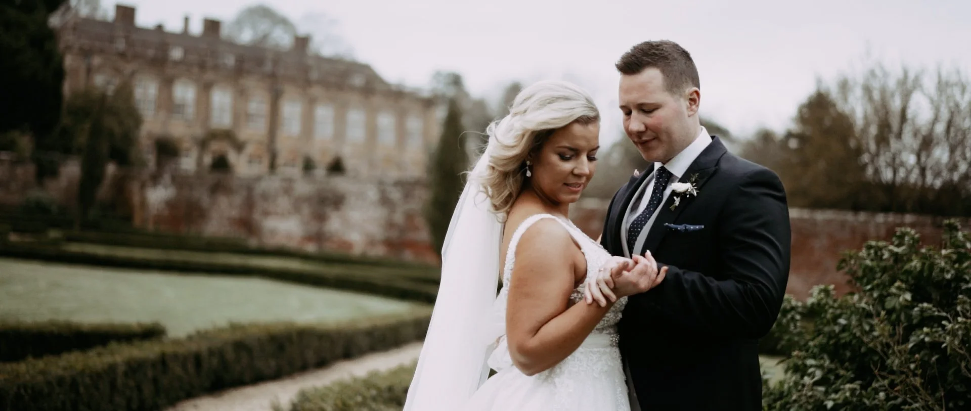 A bride and groom standing closely outdoors, with the groom holding the bride's hand, during a wedding photo shoot. The bride has blonde hair and is wearing a white wedding dress, and the groom is in a dark suit with a boutonniere. In the background, there is a historic building and trees.