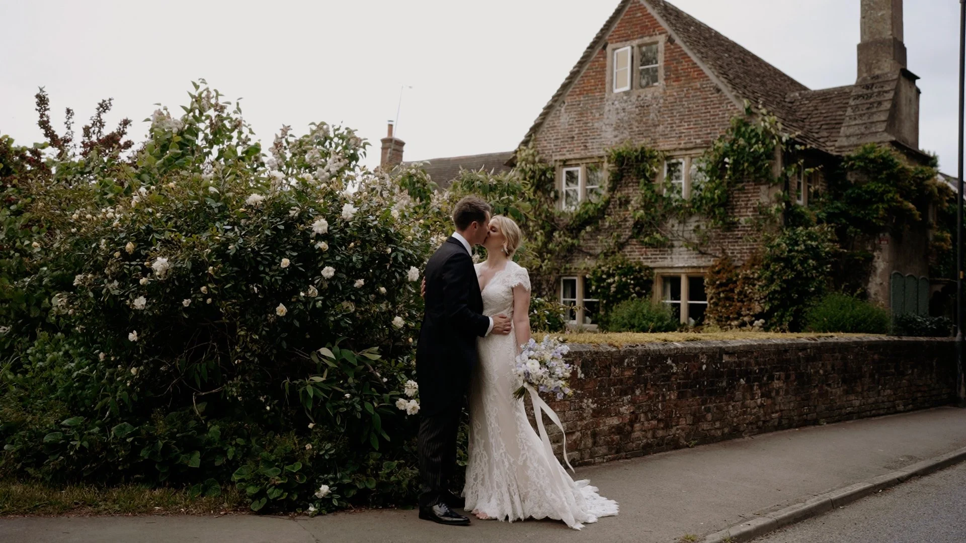 A bride and groom kiss outdoors near a brick wall and a blooming bush on a cloudy day. The bride is holding a bouquet of flowers and is wearing a lace wedding gown, while the groom is dressed in a black tuxedo.