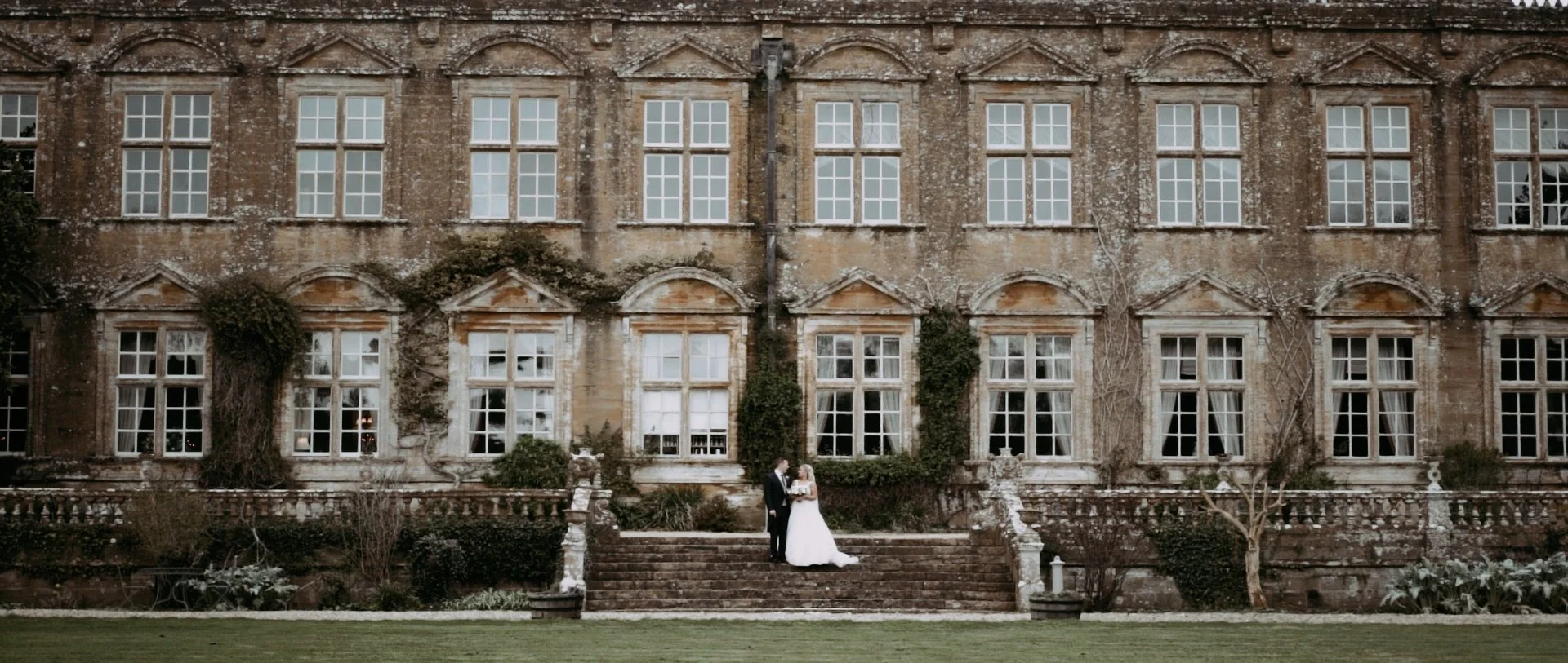 A bride and groom standing on stone steps in front of an old brick building with large windows, ivy, and garden plants.
