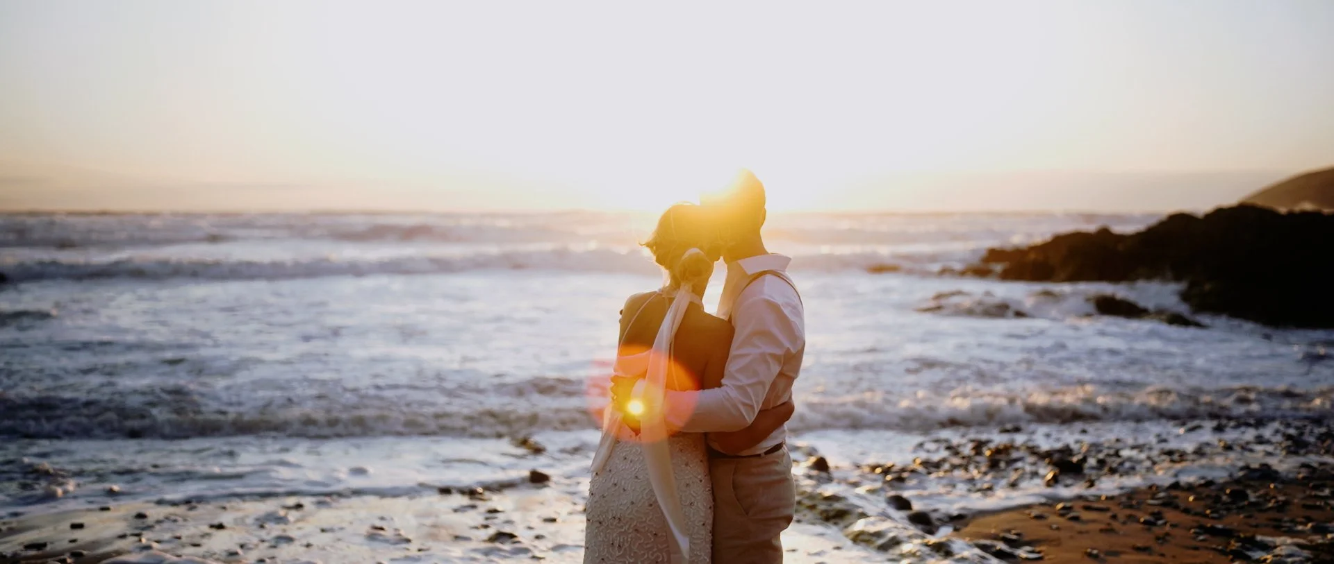 A couple dressed in wedding attire embracing on a beach at sunset.
